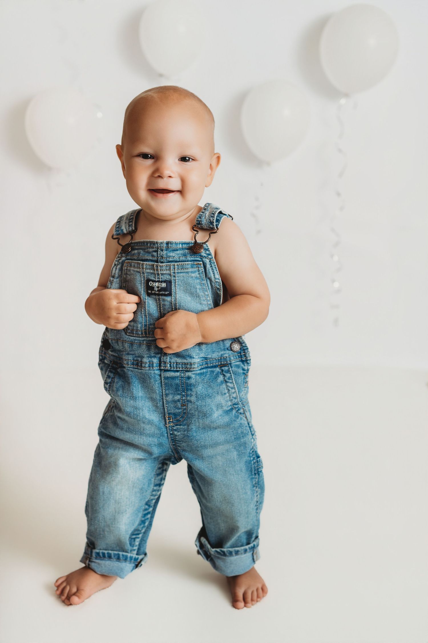 Smiling baby in denim overalls standing barefoot against white wall with white balloons.