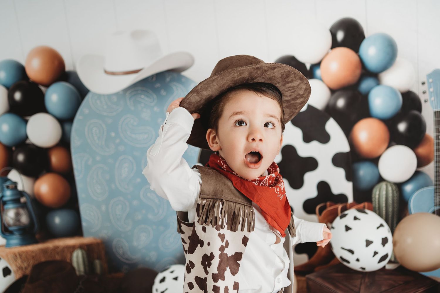 A toddler in a cowboy costume with hat and bandana poses in front of colorful balloons at a western-themed party.