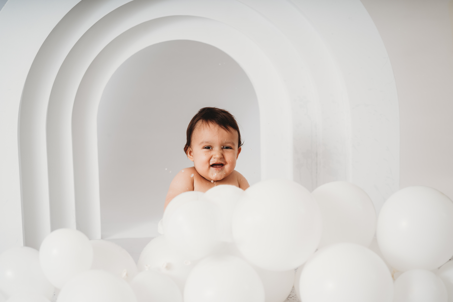 Happy baby surrounded by white balloons in a white arch backdrop studio photo session.