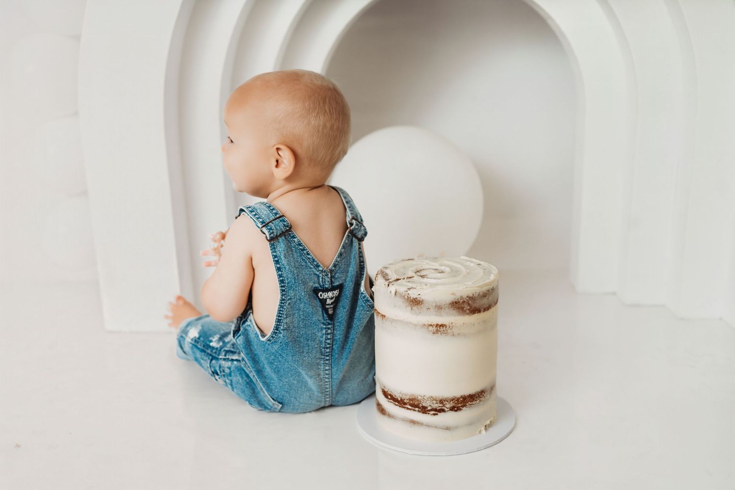 Baby in denim overalls sitting beside a naked birthday cake against a white backdrop.