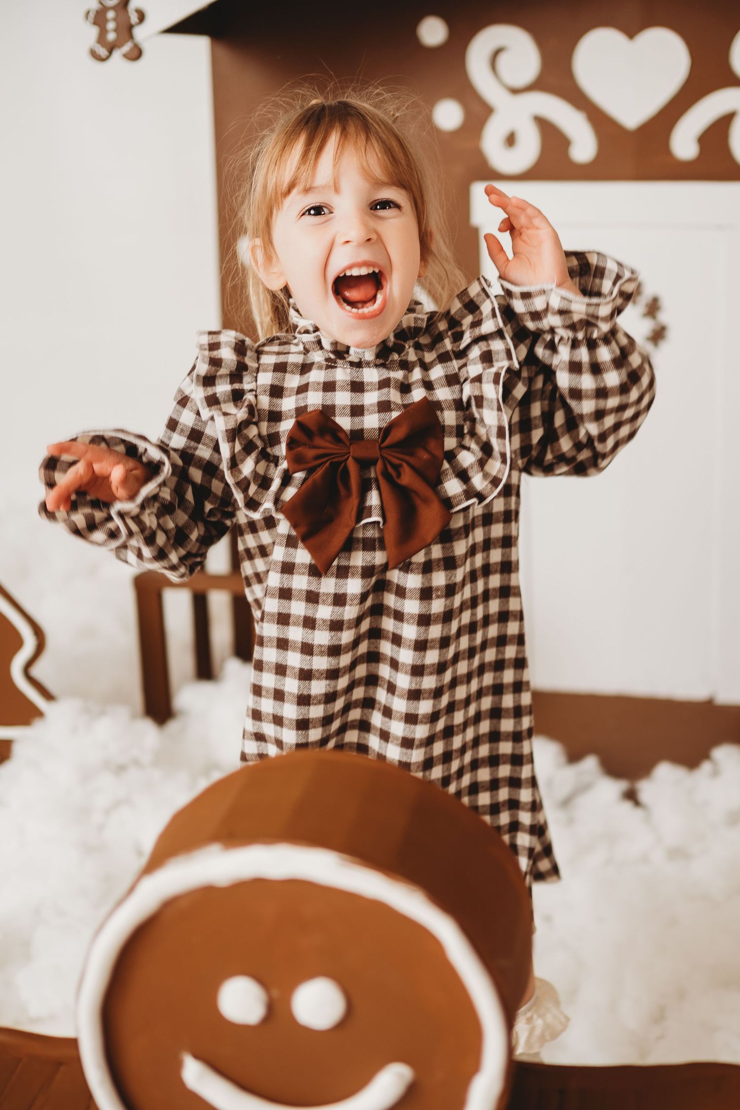 A joyful little girl in a plaid dress with bow poses excitedly near a large gingerbread man cookie decoration.