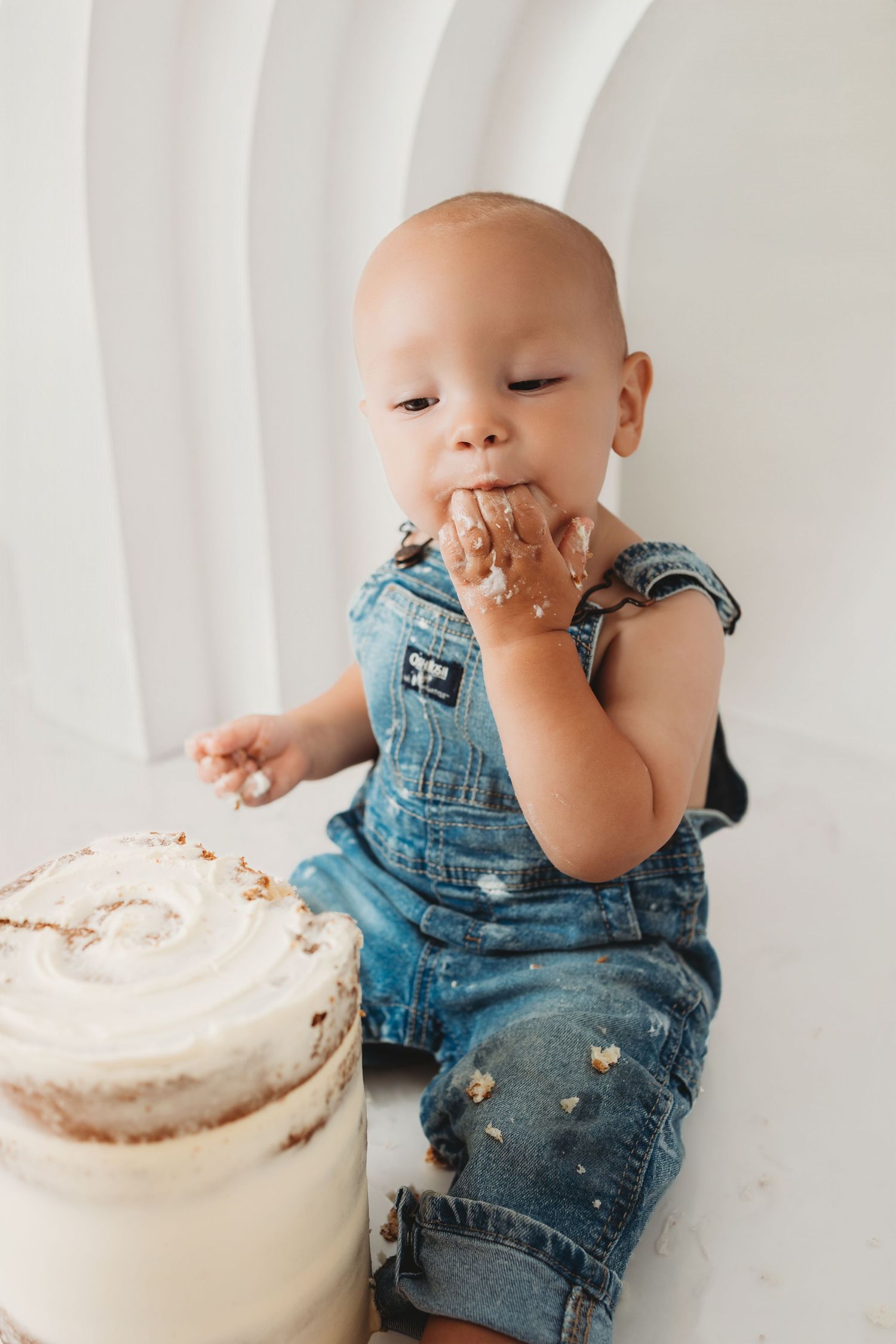 Baby in denim overalls eating cake with hands during first birthday cake smash photo session.