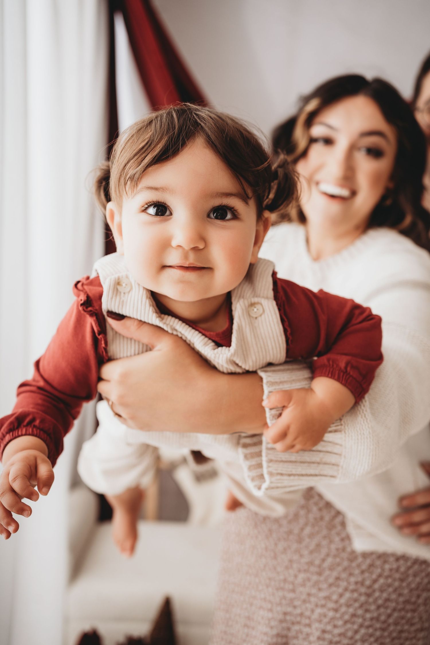 A smiling mother holds her adorable baby in a white outfit, both sharing a joyful moment indoors.