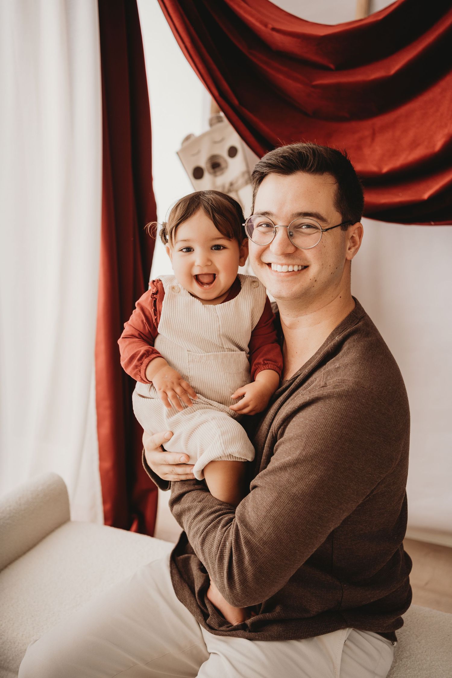 A smiling young man wearing glasses holds a laughing toddler in a bright room with red curtains.