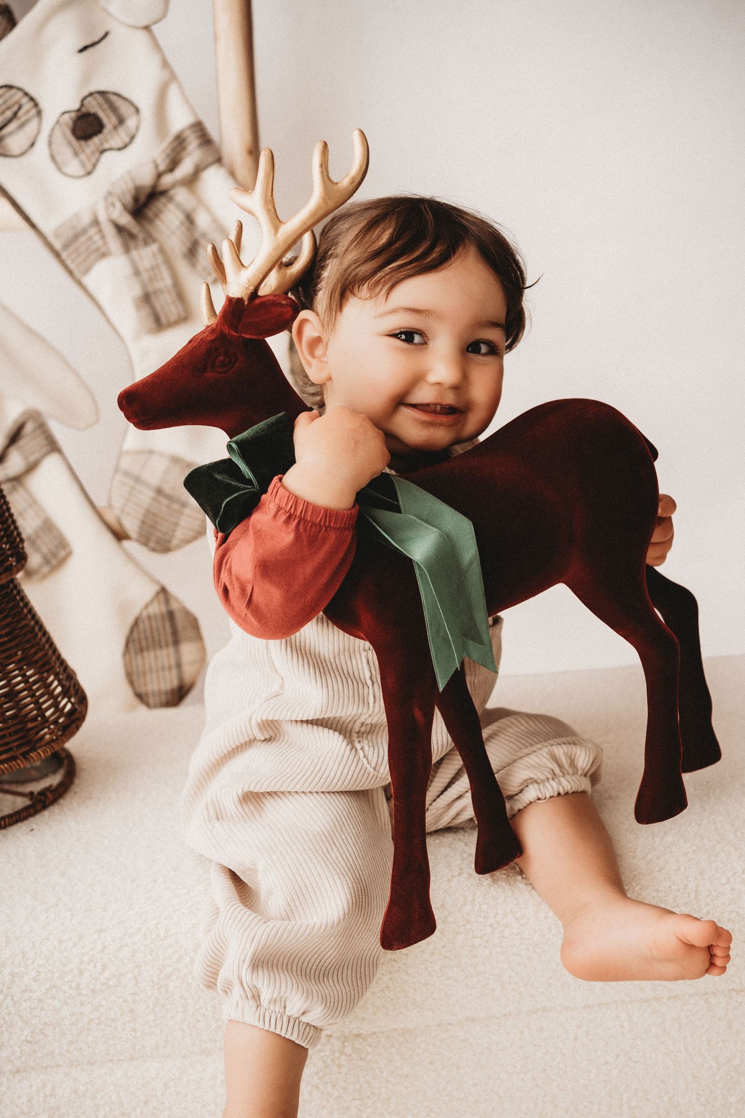 Smiling toddler in rust and cream outfit hugging a plush velvet reindeer toy on a white floor.