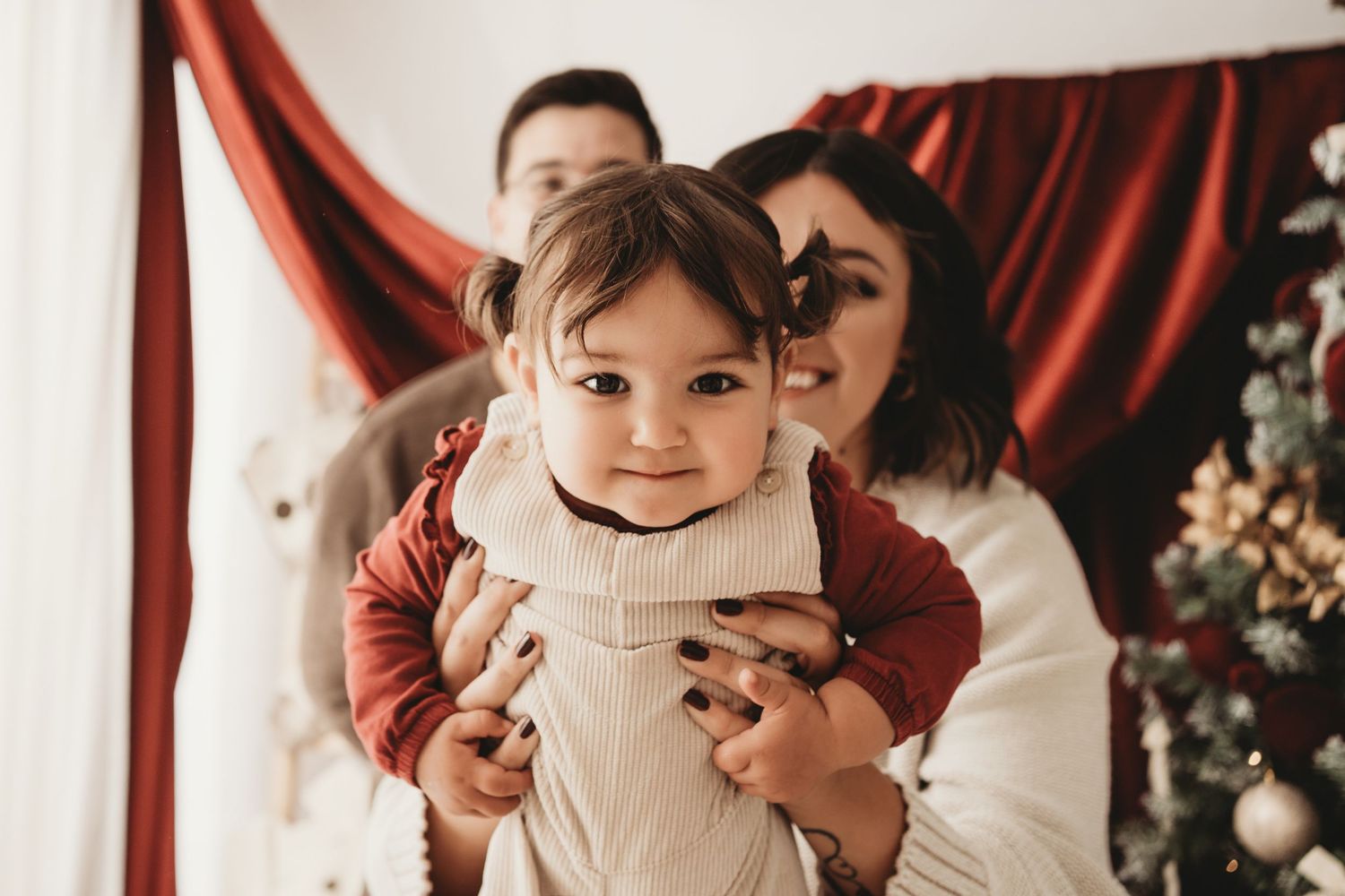 A happy baby in red outfit held by parents near a Christmas tree with festive red drapes backdrop.