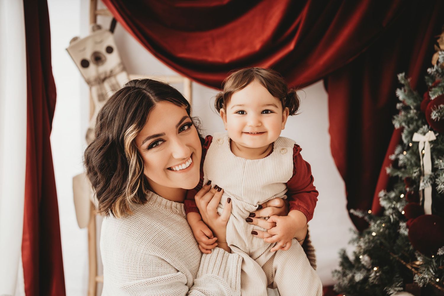 A smiling woman holds a happy toddler in a festive Christmas setting with red curtains and a decorated tree.