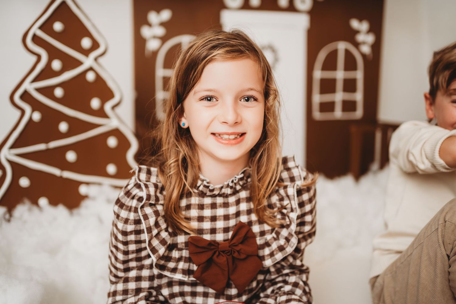 Smiling girl in plaid dress with red bow poses in front of festive gingerbread house Christmas backdrop.