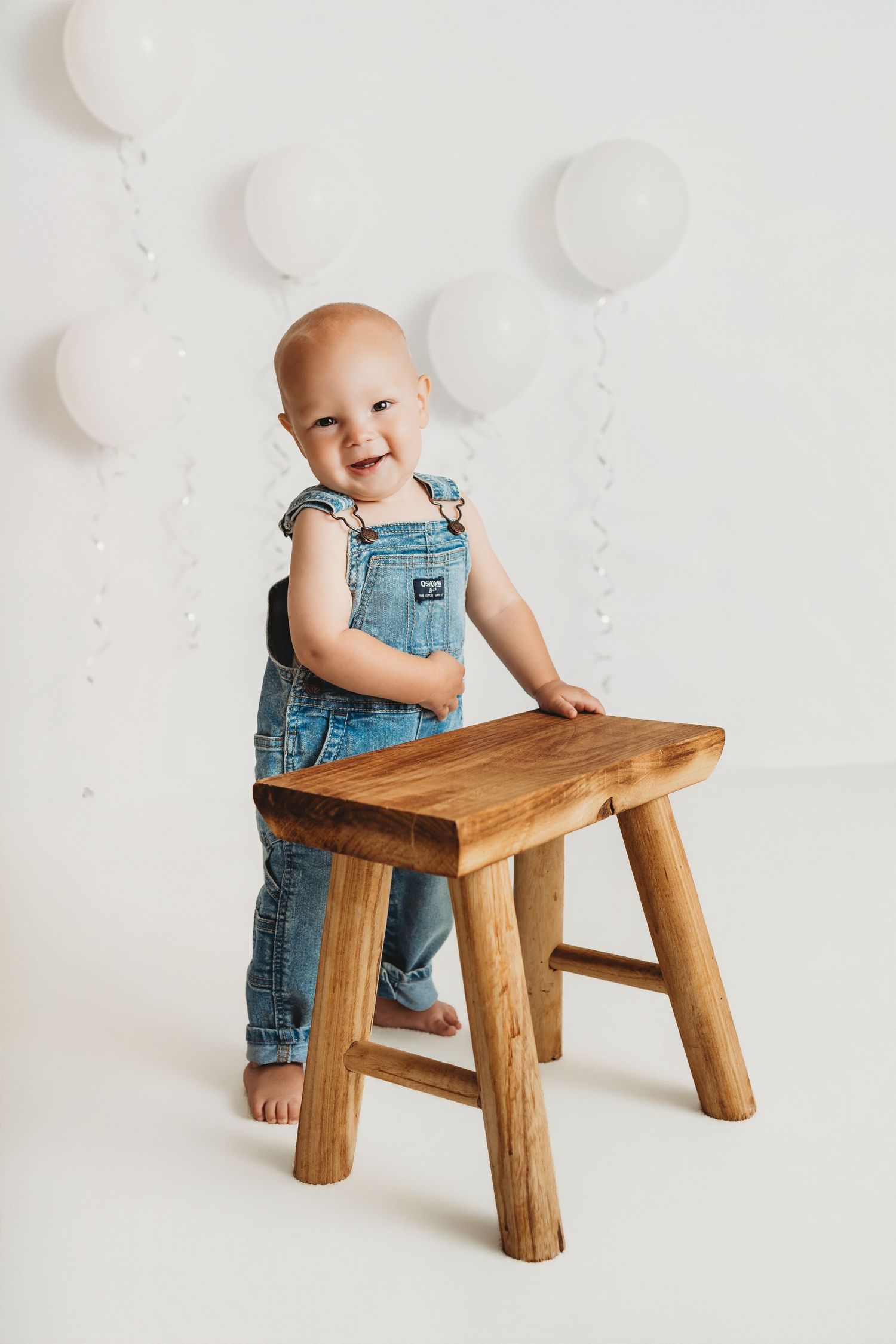 Smiling baby in denim overalls standing next to a wooden stool with white balloons in the background.