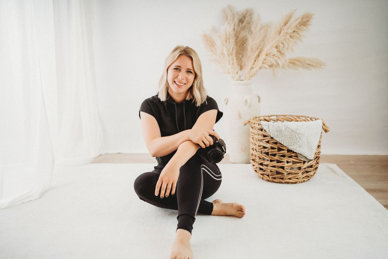 Smiling blonde woman in black athletic wear sitting cross-legged on white floor near wicker basket and pampas grass.