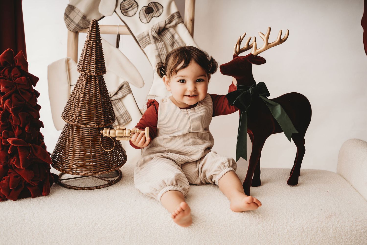 Smiling baby in beige romper sits among Christmas decorations including a reindeer figurine and wicker tree.