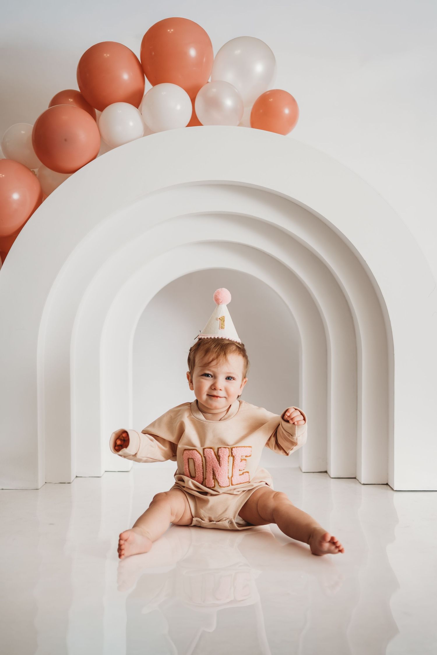 Smiling baby in ONE outfit and party hat sits before white rainbow arches with pink and white balloons.
