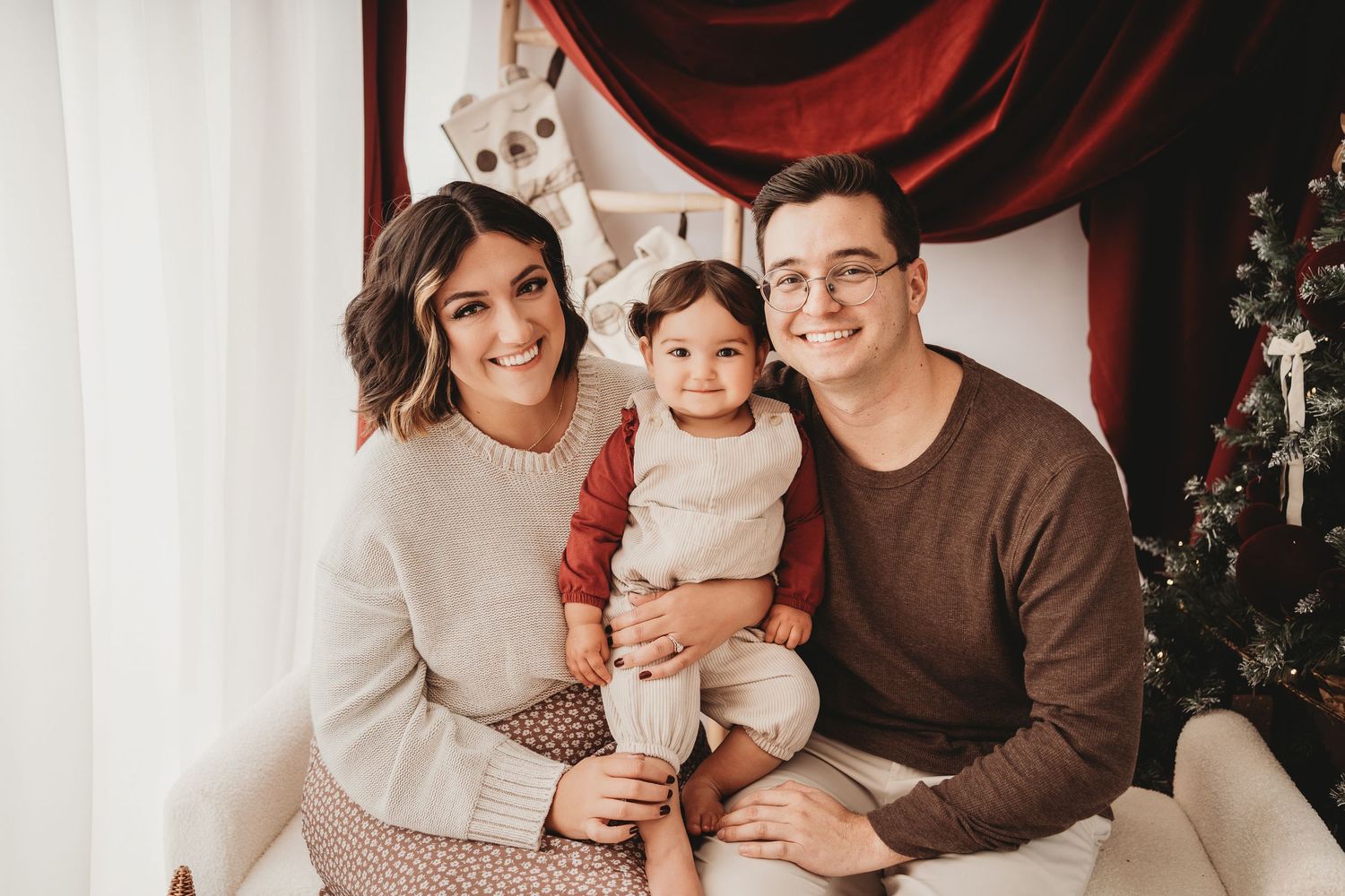 Family of three posing for a Christmas photo, parents smiling with toddler in holiday setting.