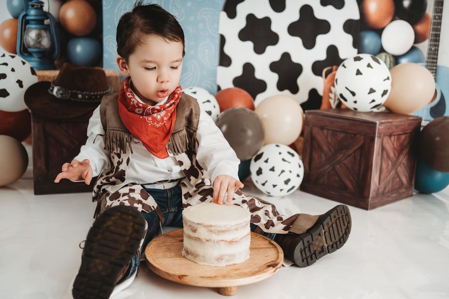 Toddler boy in cowboy outfit with bandana sits beside smash cake, surrounded by balloons and wooden crates.