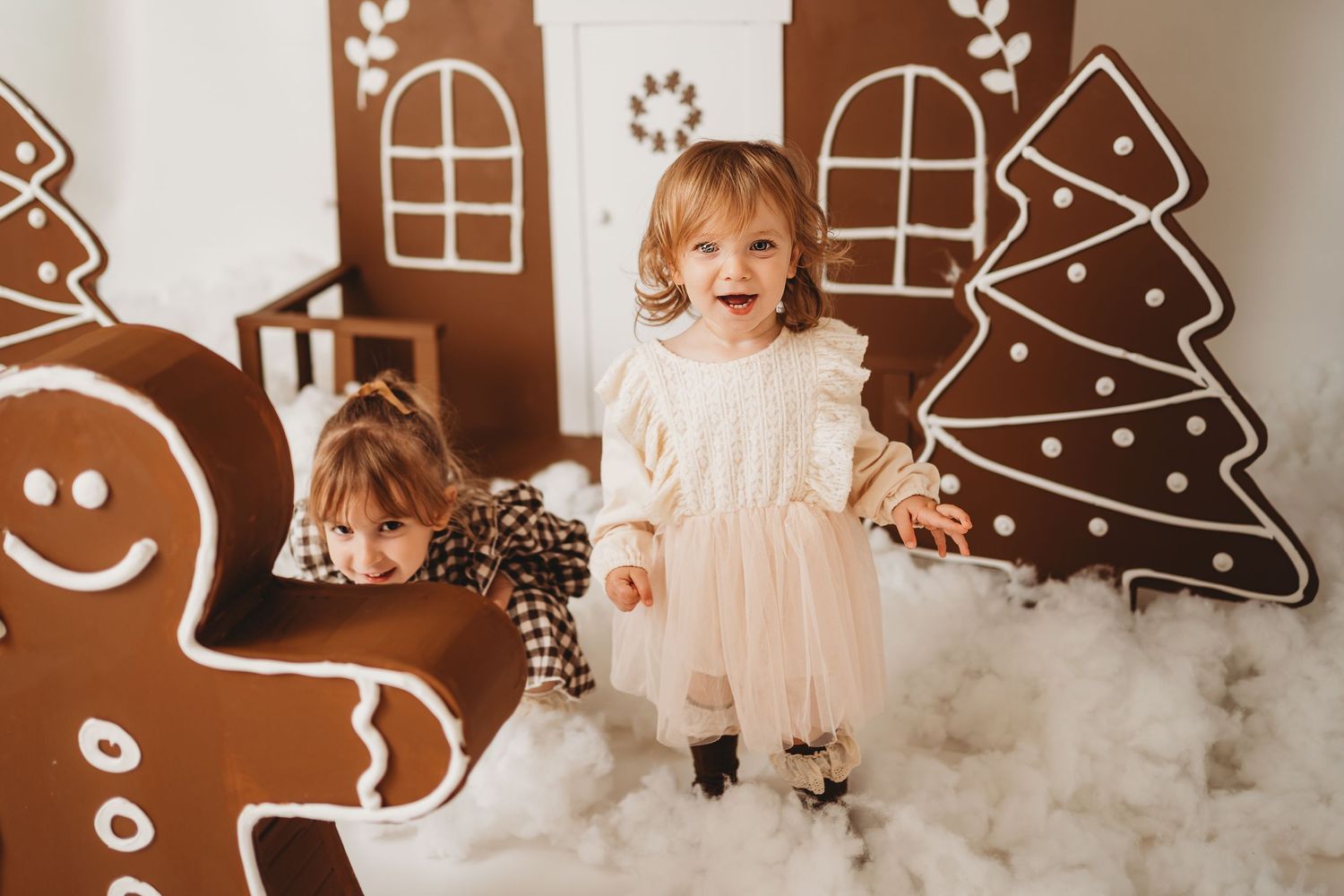 Two young children pose joyfully in a festive Christmas gingerbread house photo set with decorative trees.
