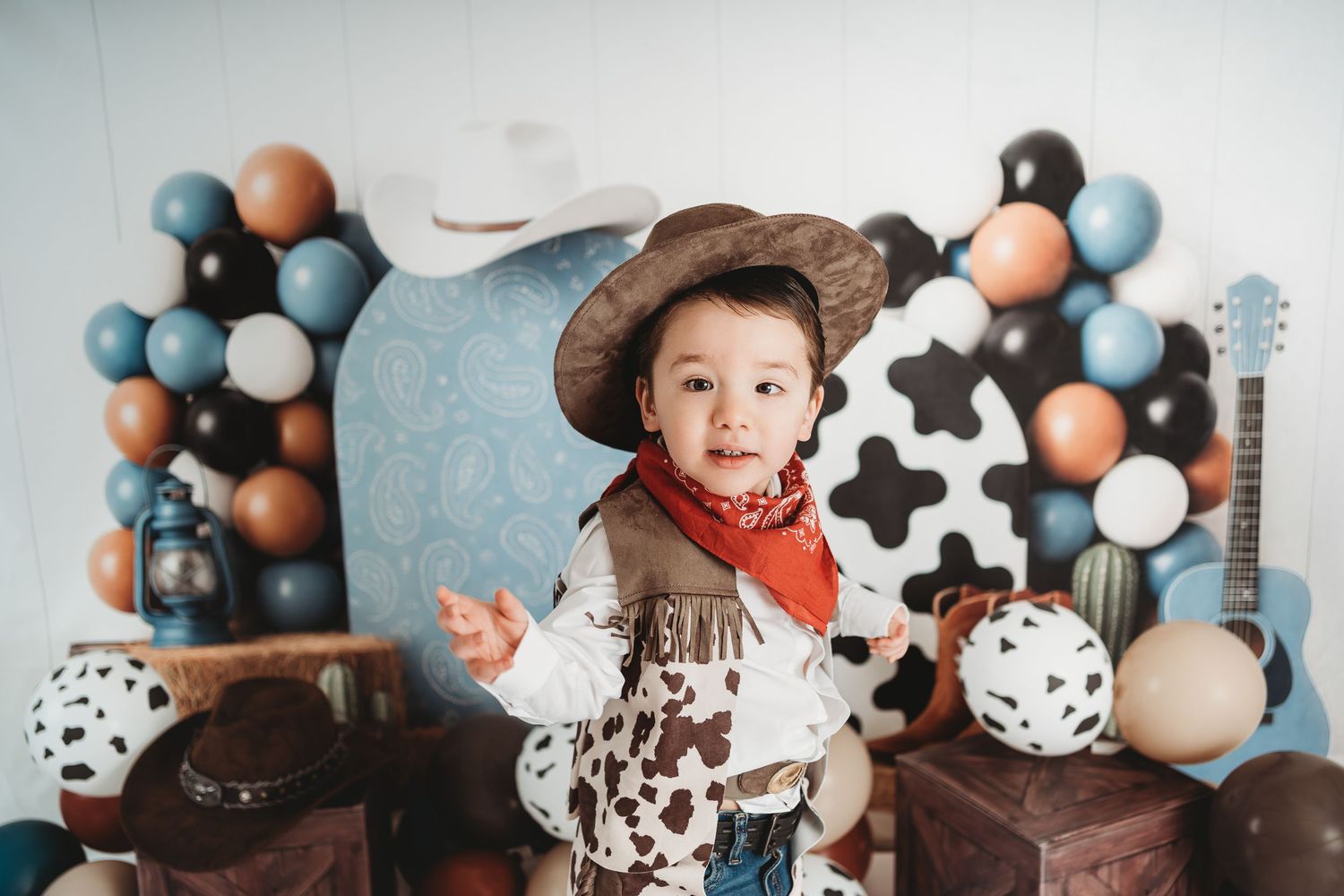Toddler in cowboy hat and vest poses at a Western-themed birthday party with balloons and decorations.