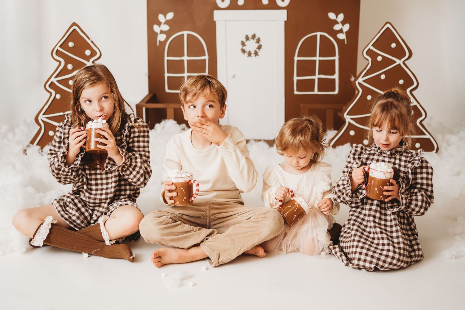 Four children in matching plaid pajamas sit together drinking hot cocoa in front of a gingerbread house Christmas backdrop.