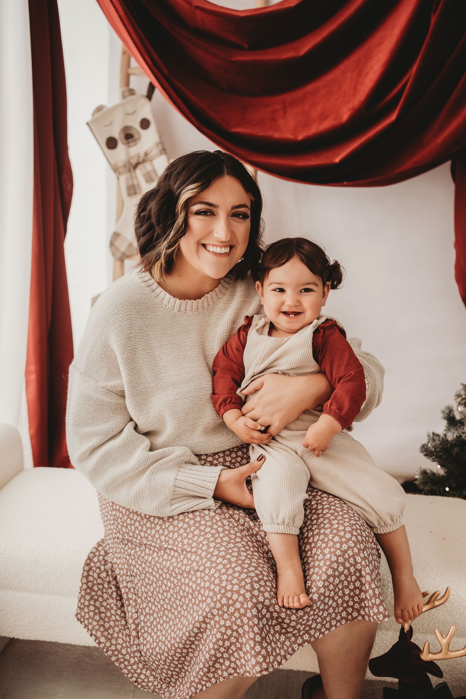 A smiling mother holds her happy toddler during a festive Christmas photo session with red drapes.
