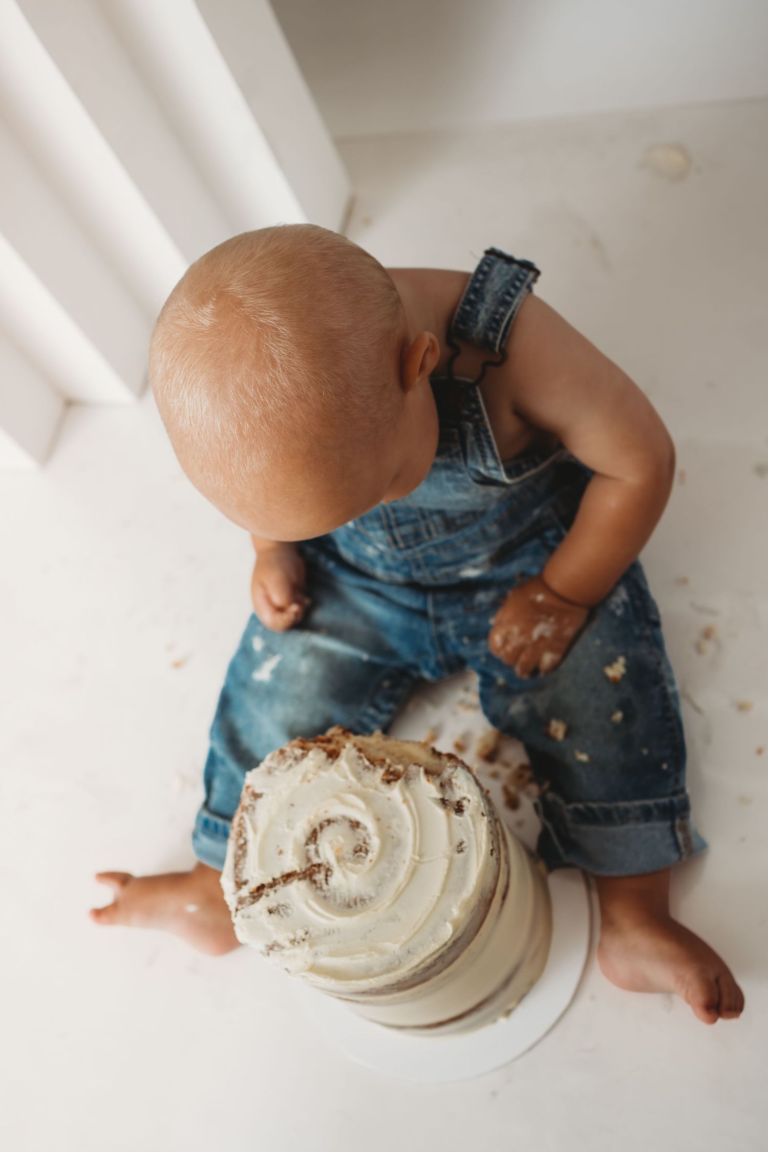 Baby in denim overalls sits on floor smashing a white frosted cake during a first birthday cake smash photo session.