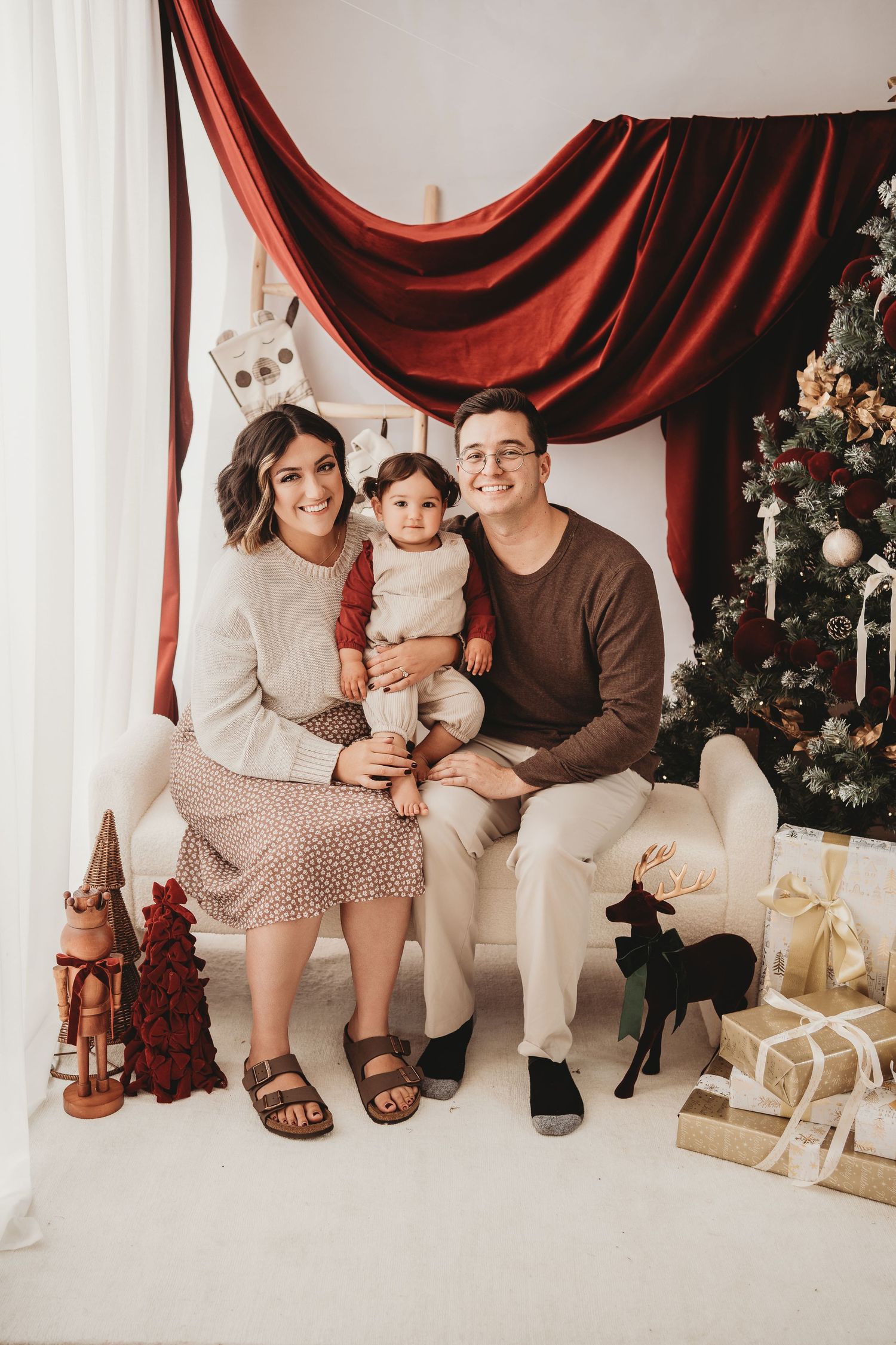Happy family of three poses for a Christmas portrait with decorated tree, gifts, and festive red velvet backdrop.
