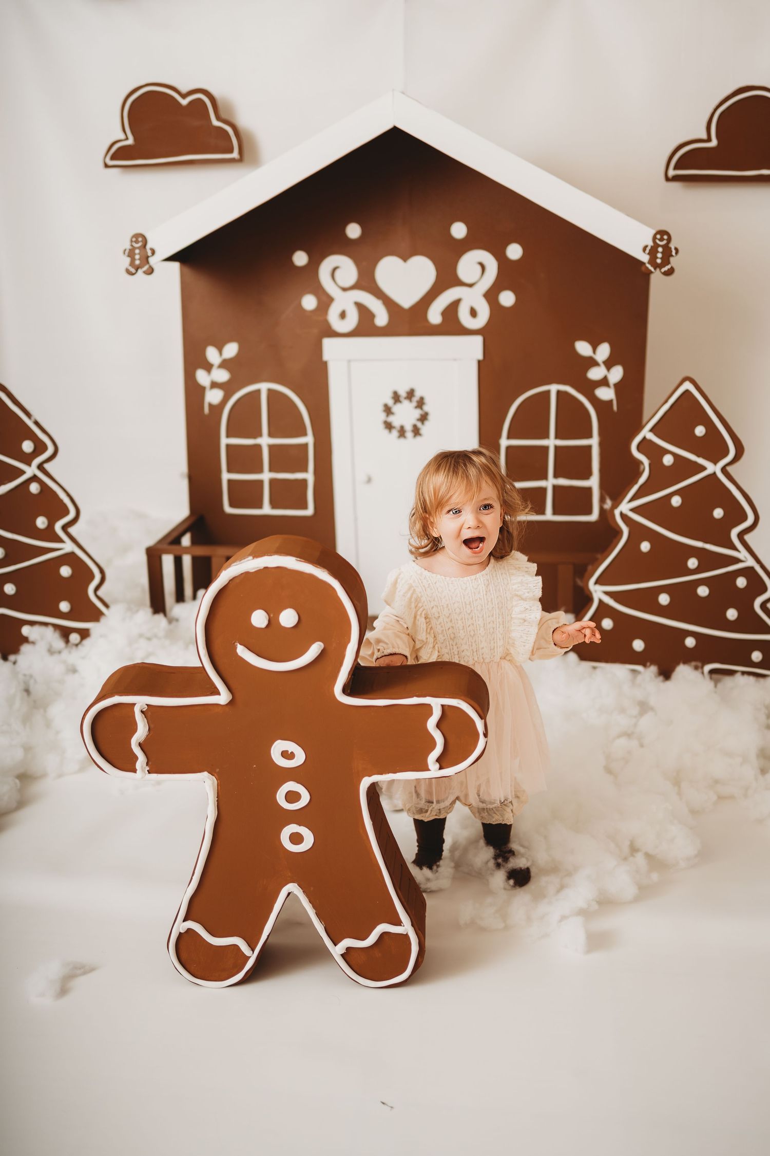 A joyful toddler girl in a white dress poses with a giant gingerbread man decoration in a festive Christmas scene.