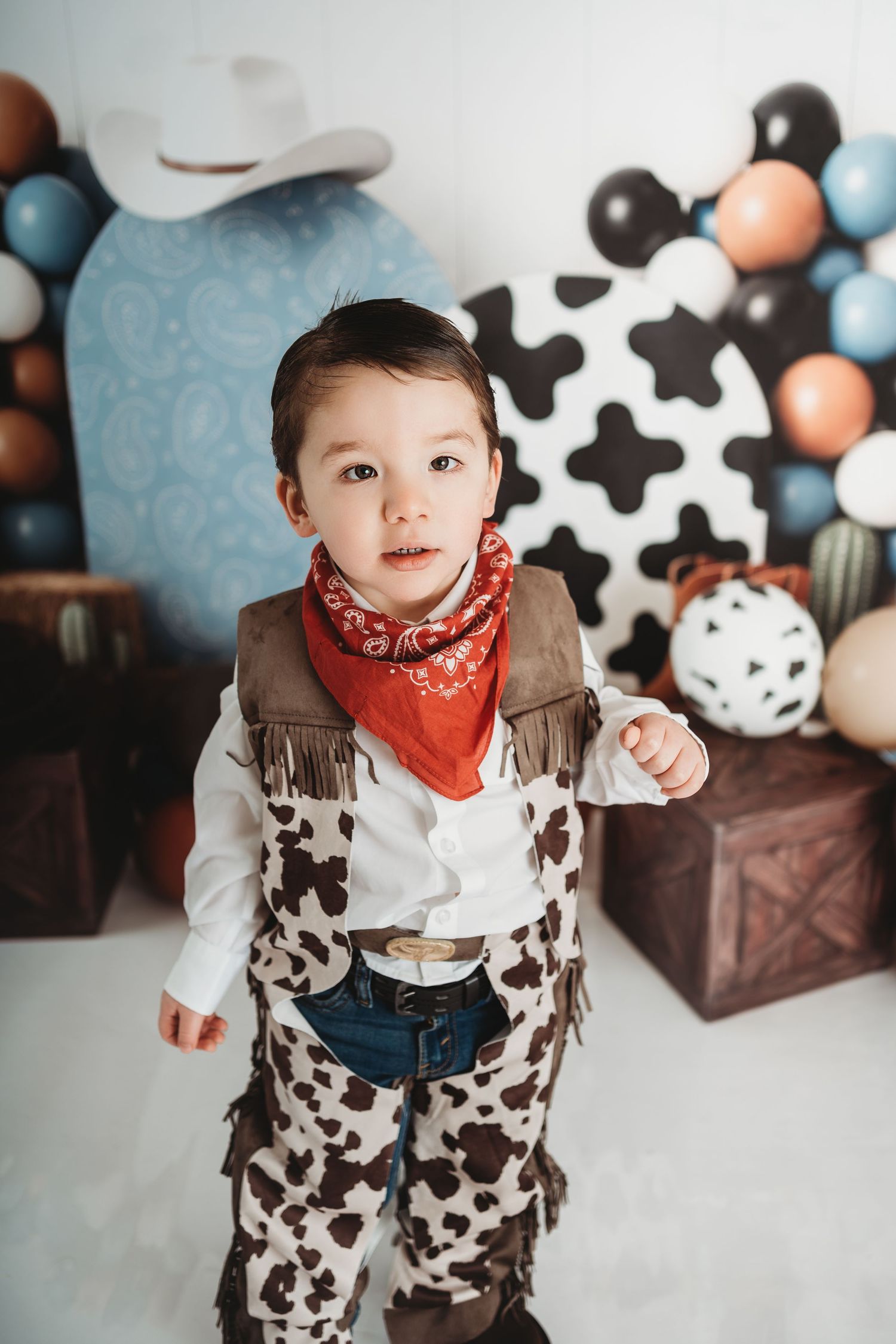 Toddler boy in cowboy costume with red bandana, fringe vest, and cow print chaps at a western themed birthday party.