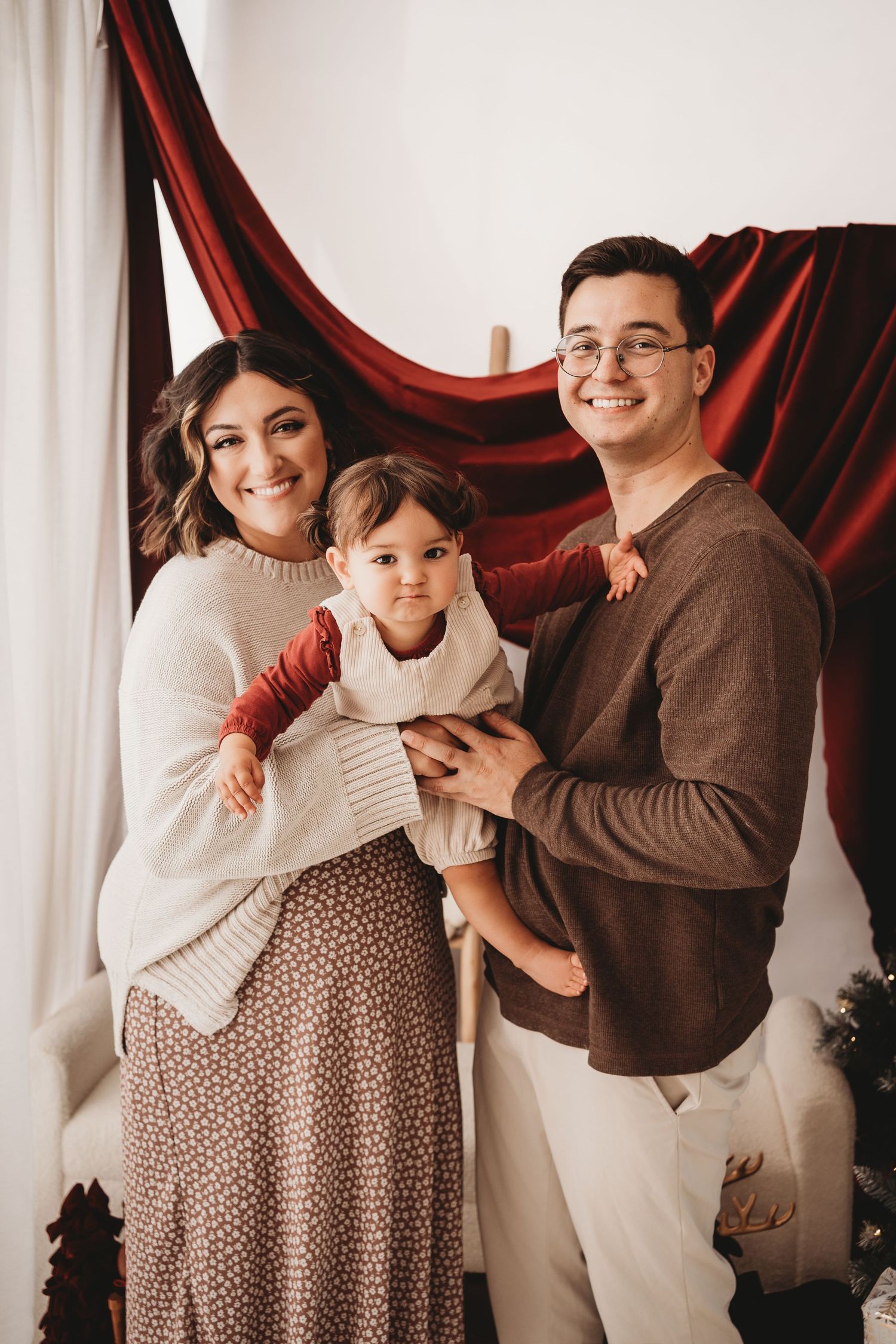 A smiling family of three poses for a holiday portrait with red draped fabric backdrop, parents holding their toddler.