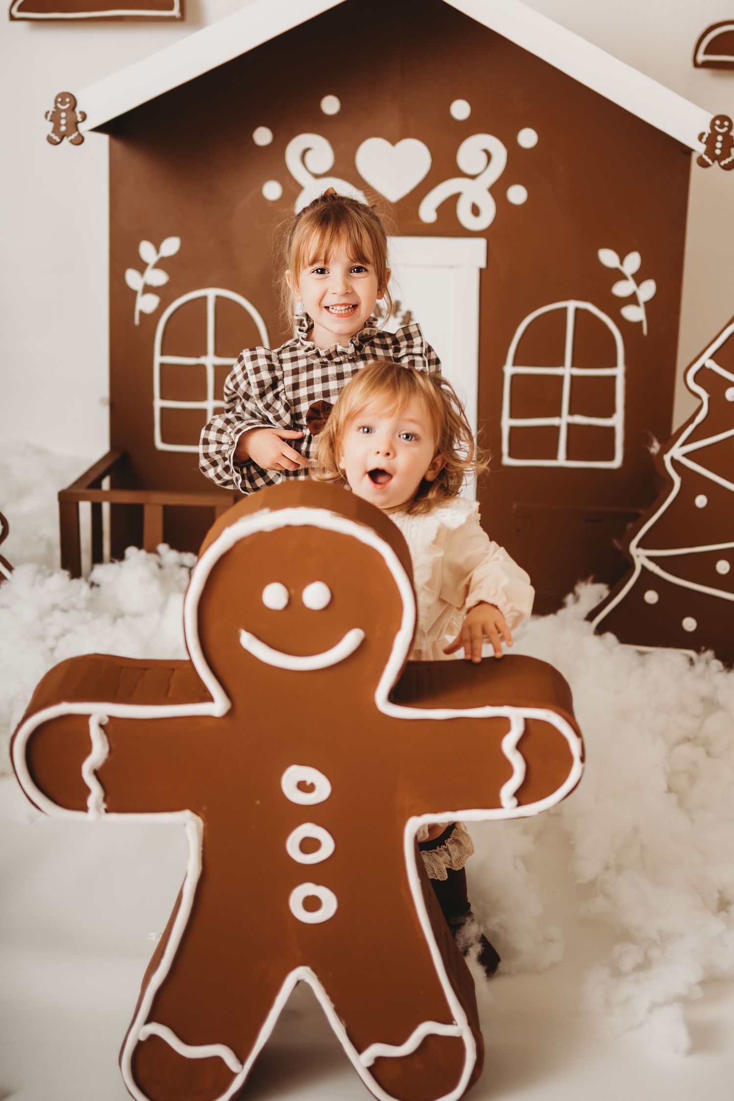 Two cheerful children pose with a giant gingerbread man cookie in front of a decorative chocolate gingerbread house.