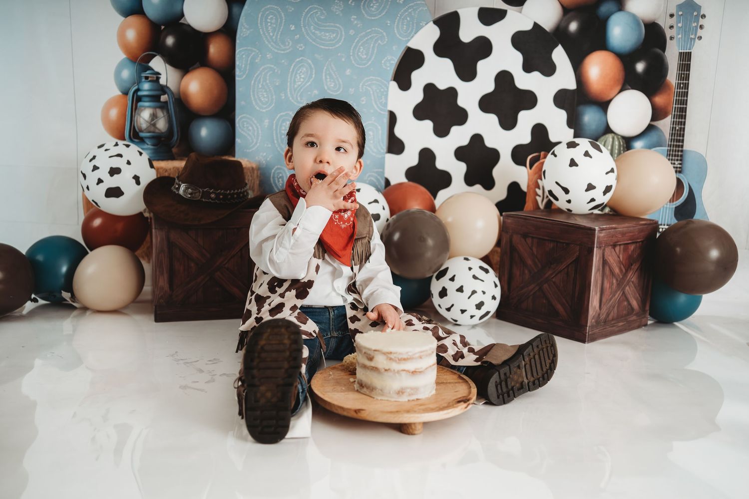 Baby boy in cowboy outfit enjoys first birthday cake smash with balloons and western-themed decorations.
