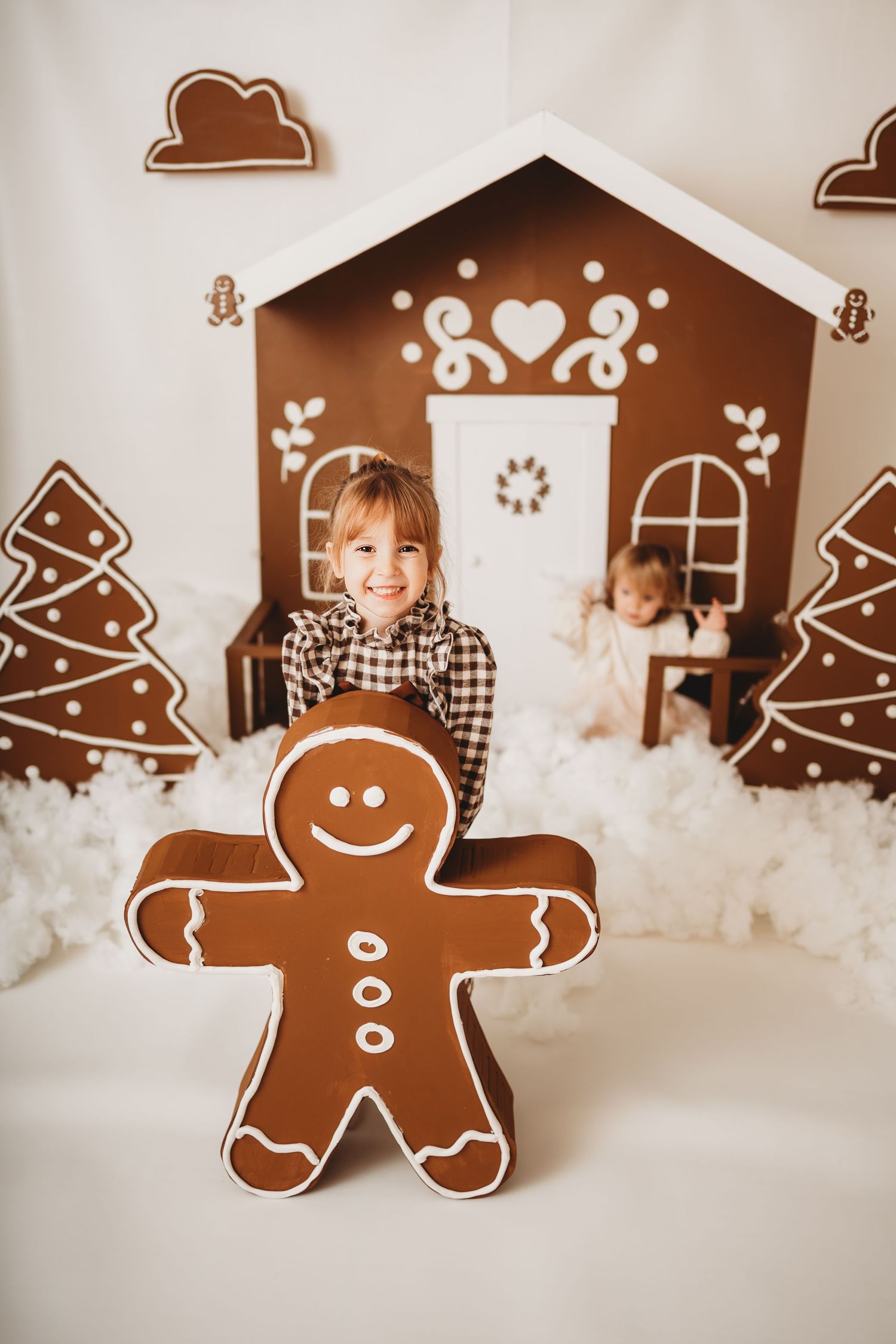 Child smiling with giant gingerbread man cookie in front of decorative gingerbread house Christmas backdrop.