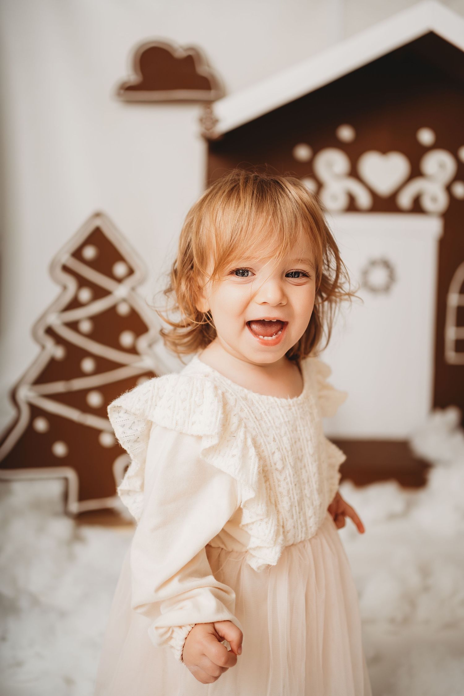 Joyful toddler girl in white dress laughing before gingerbread house Christmas decorations.