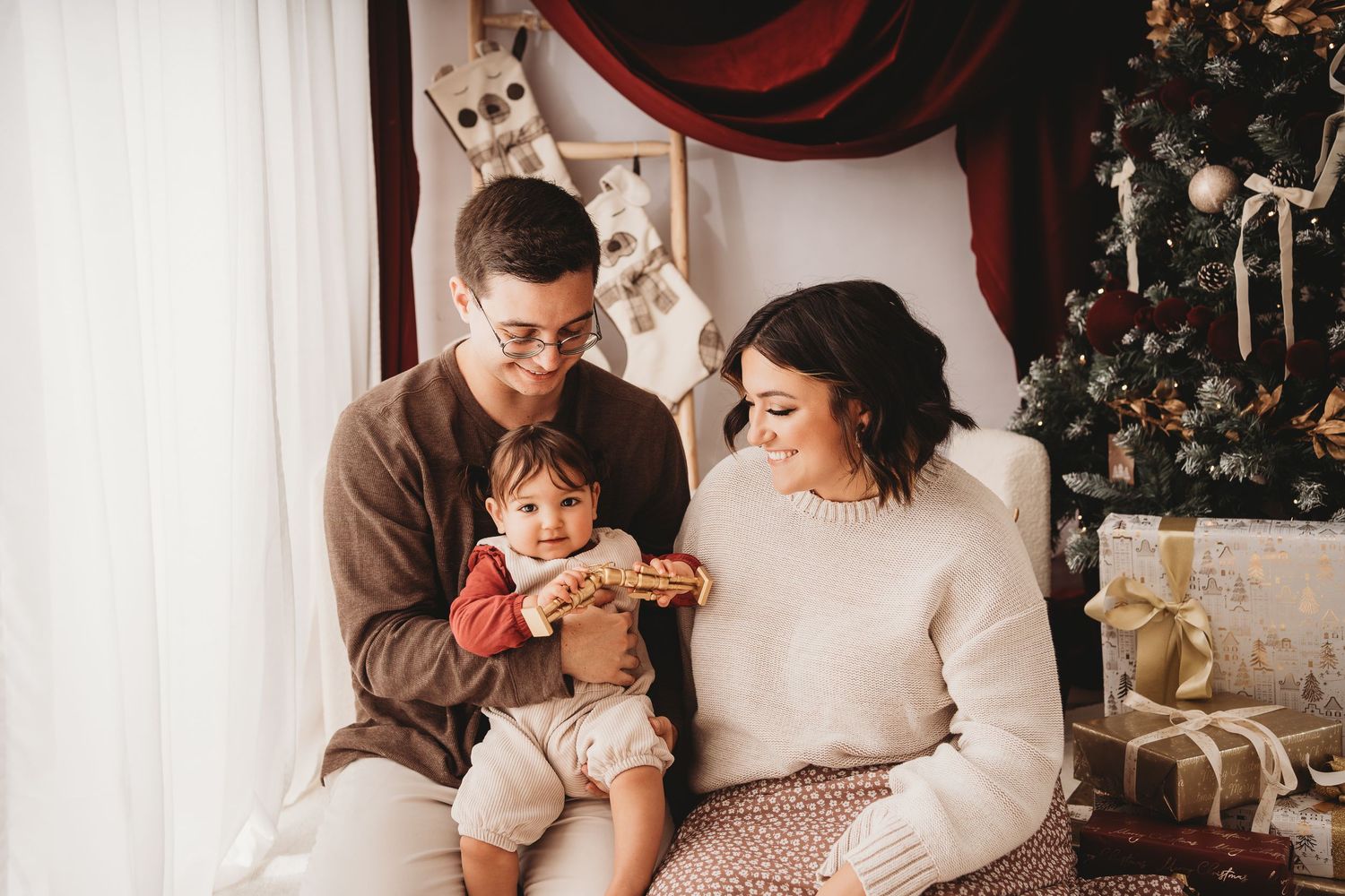 Young family with baby celebrating Christmas near decorated tree with gifts in cozy holiday setting.