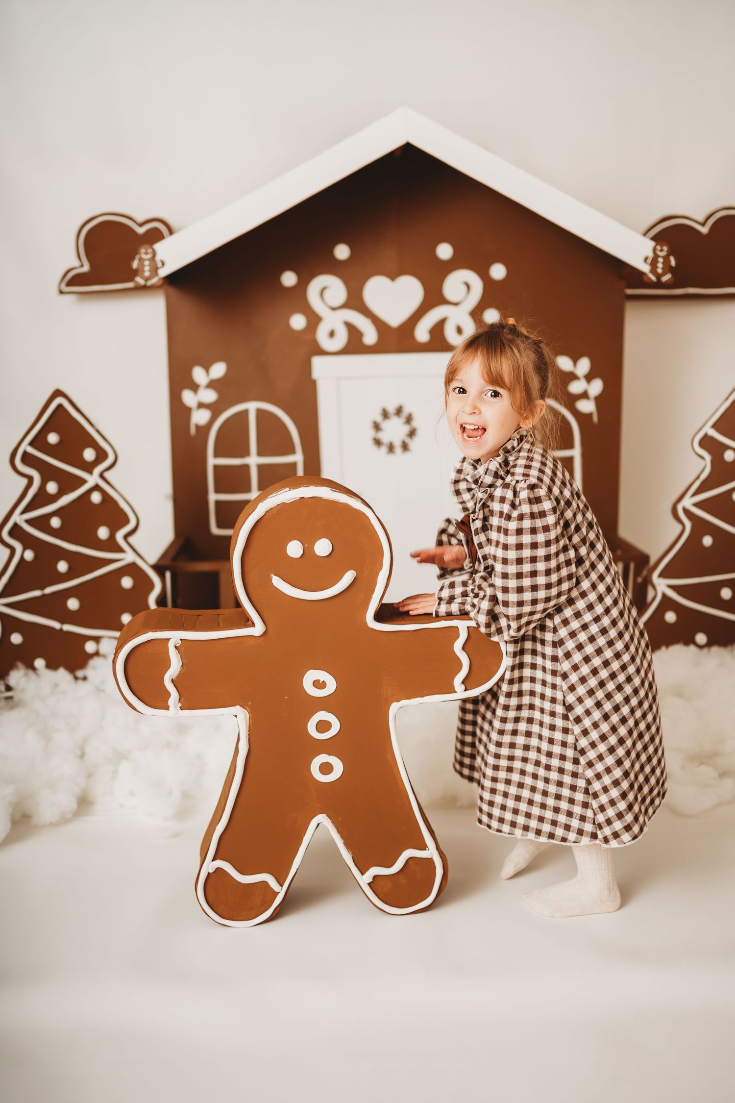 Young girl in plaid dress posing with giant gingerbread man cutout beside decorative Christmas gingerbread house backdrop.