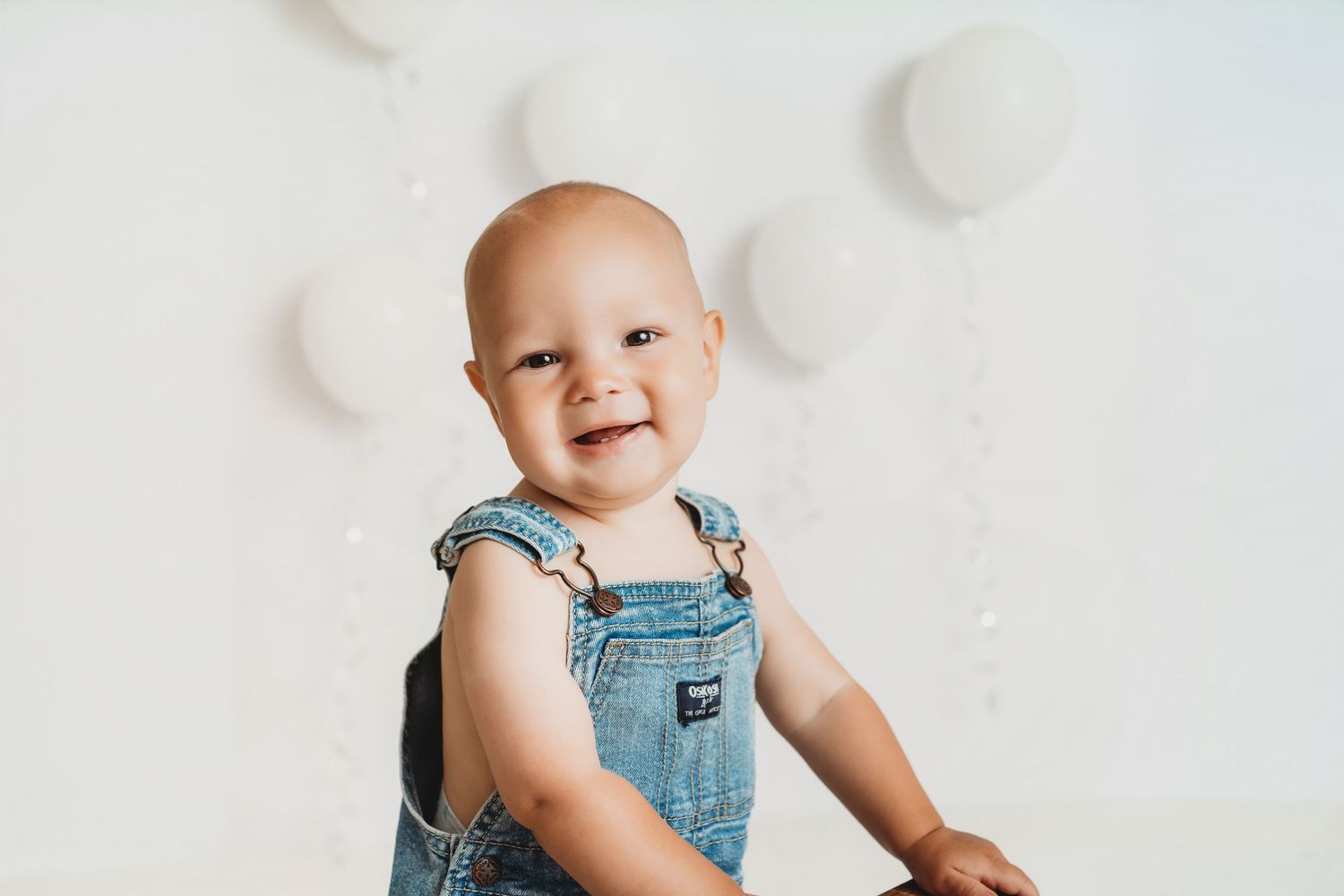 Smiling baby in denim overalls sitting in front of white balloon backdrop in a bright studio setting.