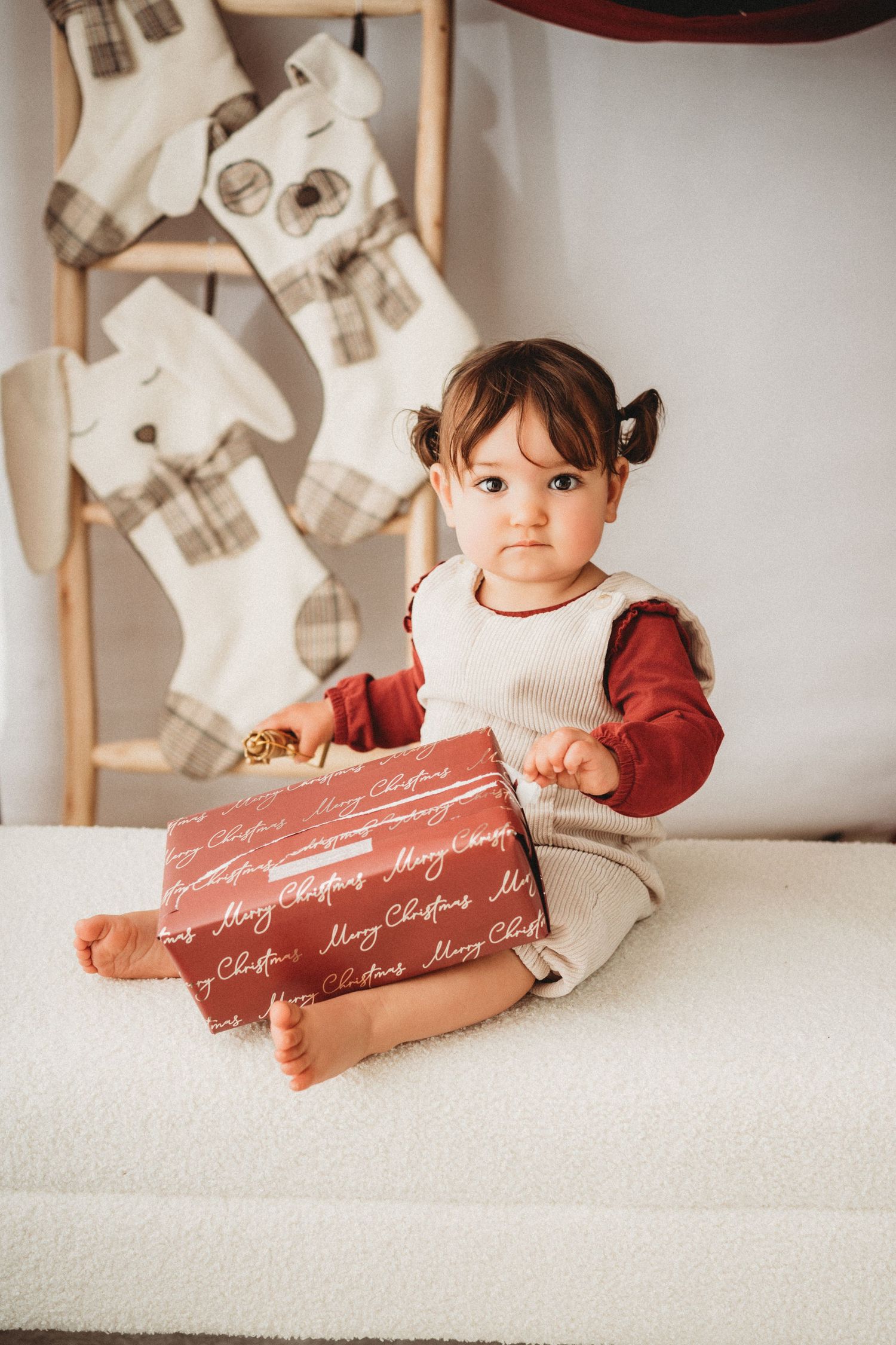 Cute baby girl in red and white outfit holding a Merry Christmas gift box near holiday stockings.