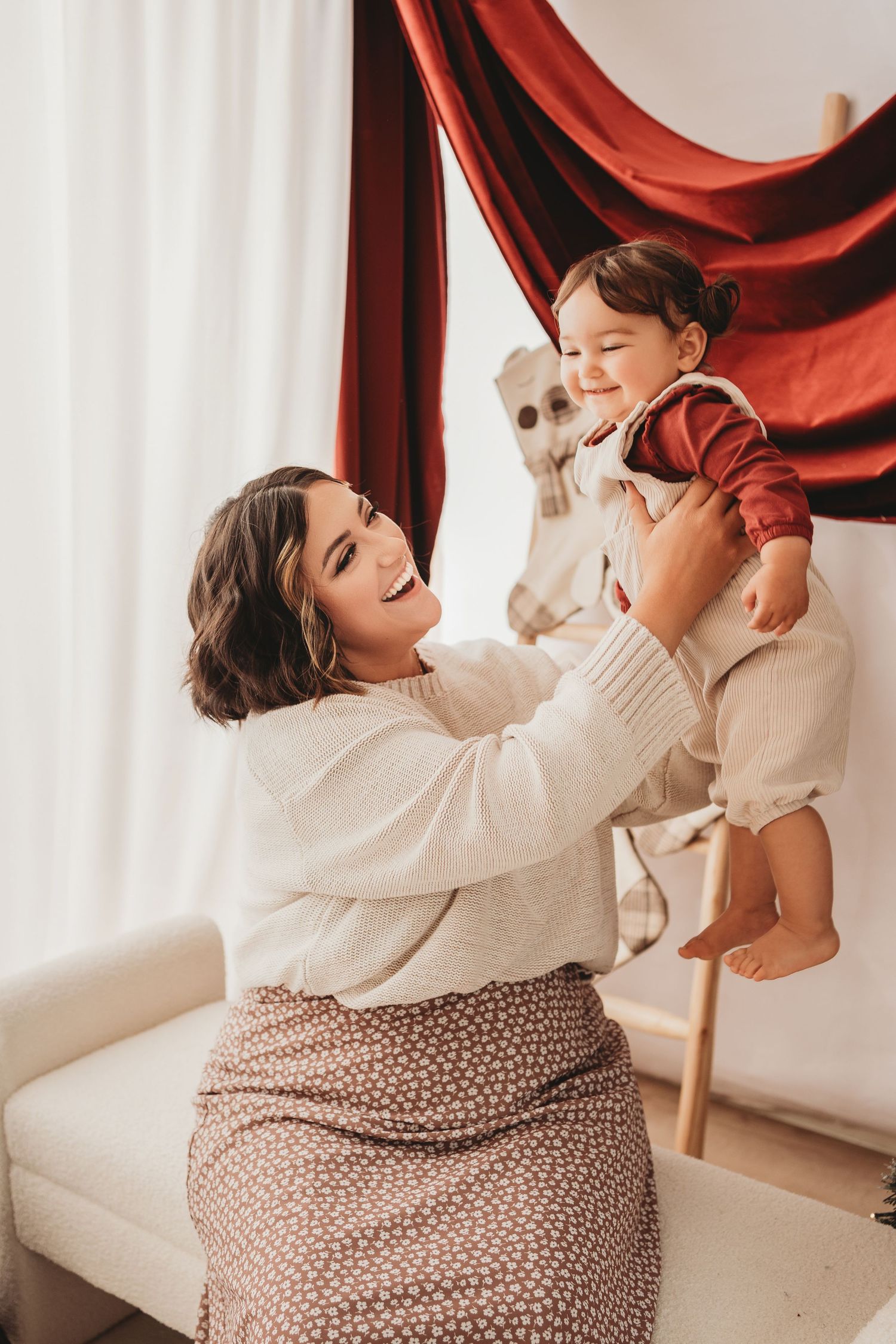 A smiling mother lifts her happy toddler in a bright room with red drape backdrop.