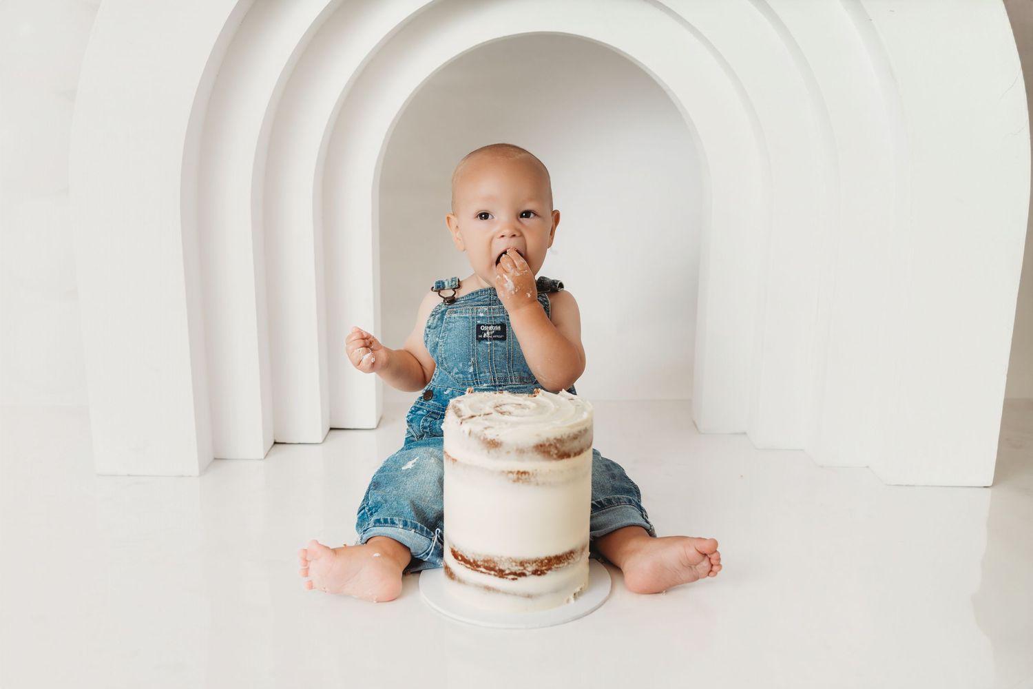 Baby in denim overalls eating cake during a first birthday smash cake photo session on white background.