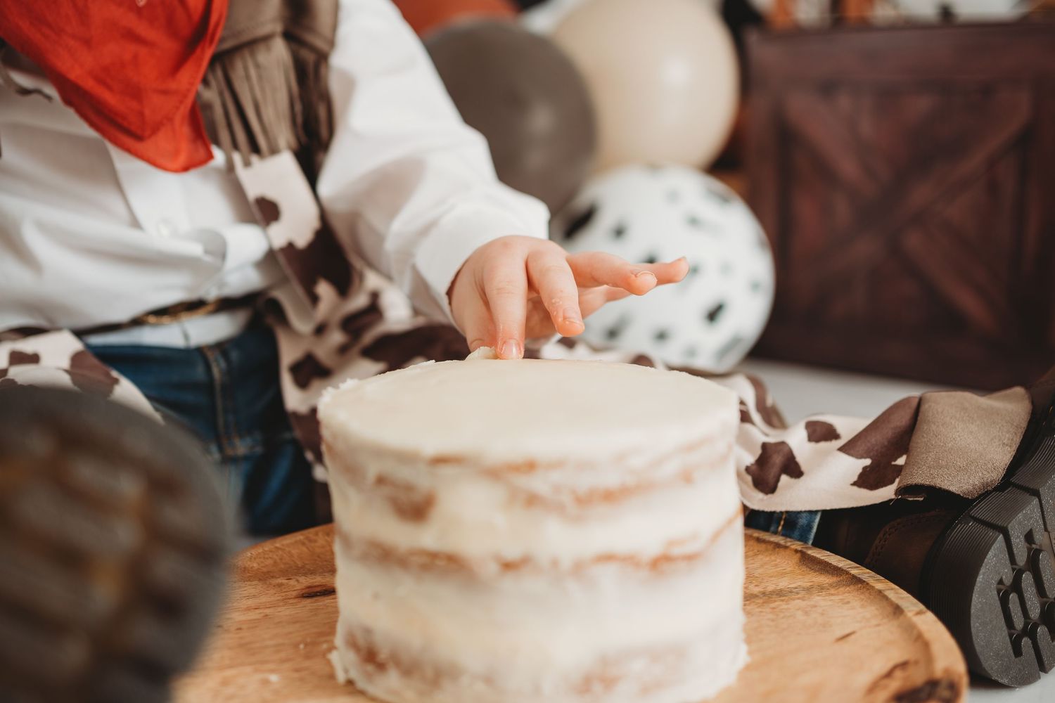 Child in cowboy outfit touching a rustic naked cake on wooden board at a western-themed birthday party.