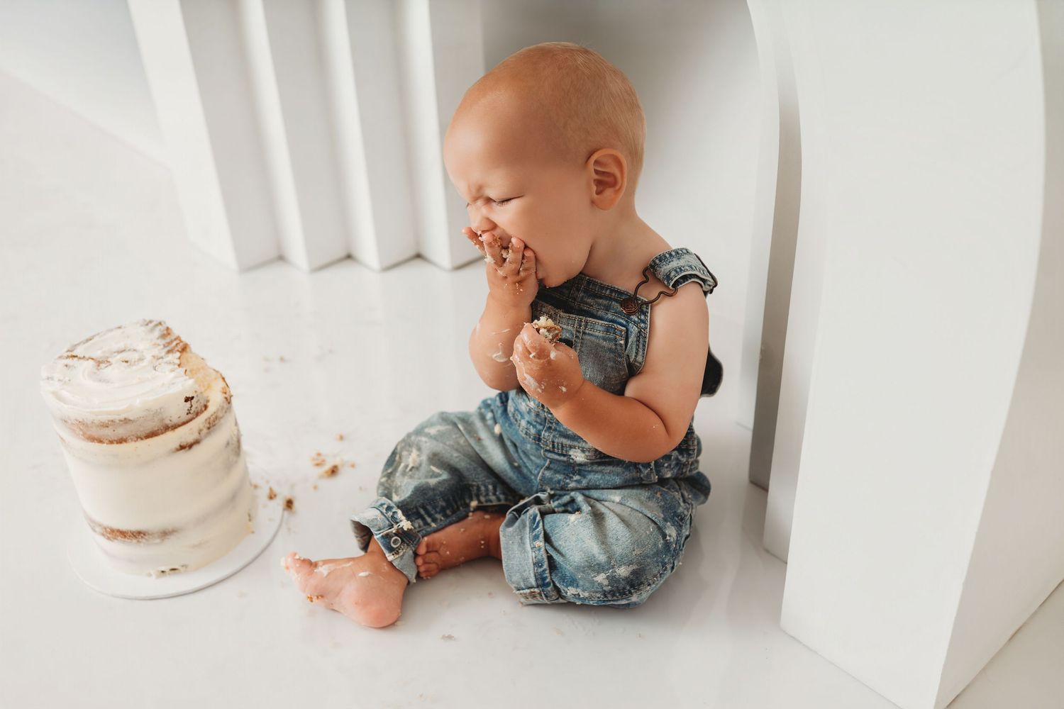 Baby in denim overalls sitting on white floor eating first birthday cake smash.