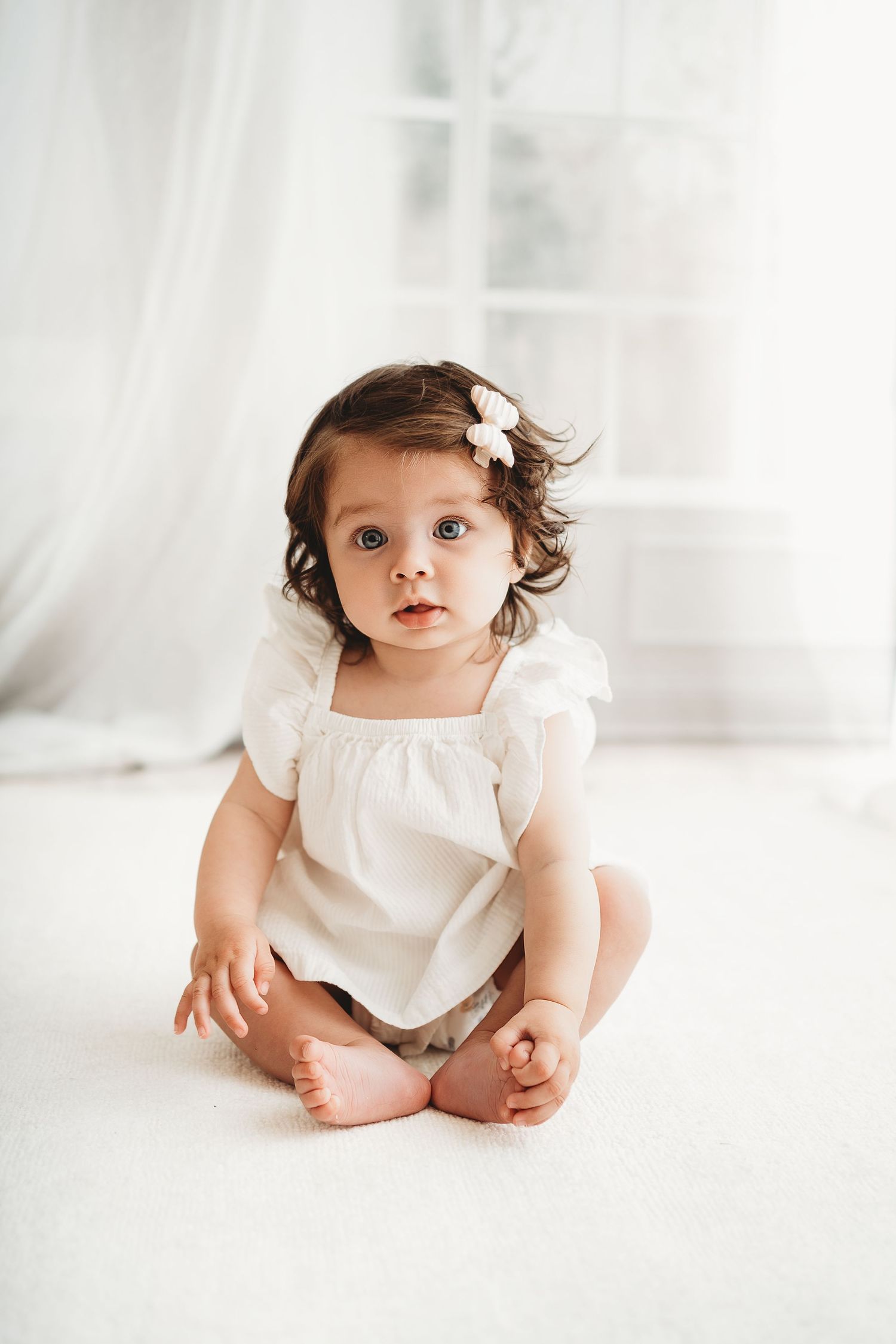 Adorable baby girl in white romper with flower hair clip sits on white floor in bright airy room.