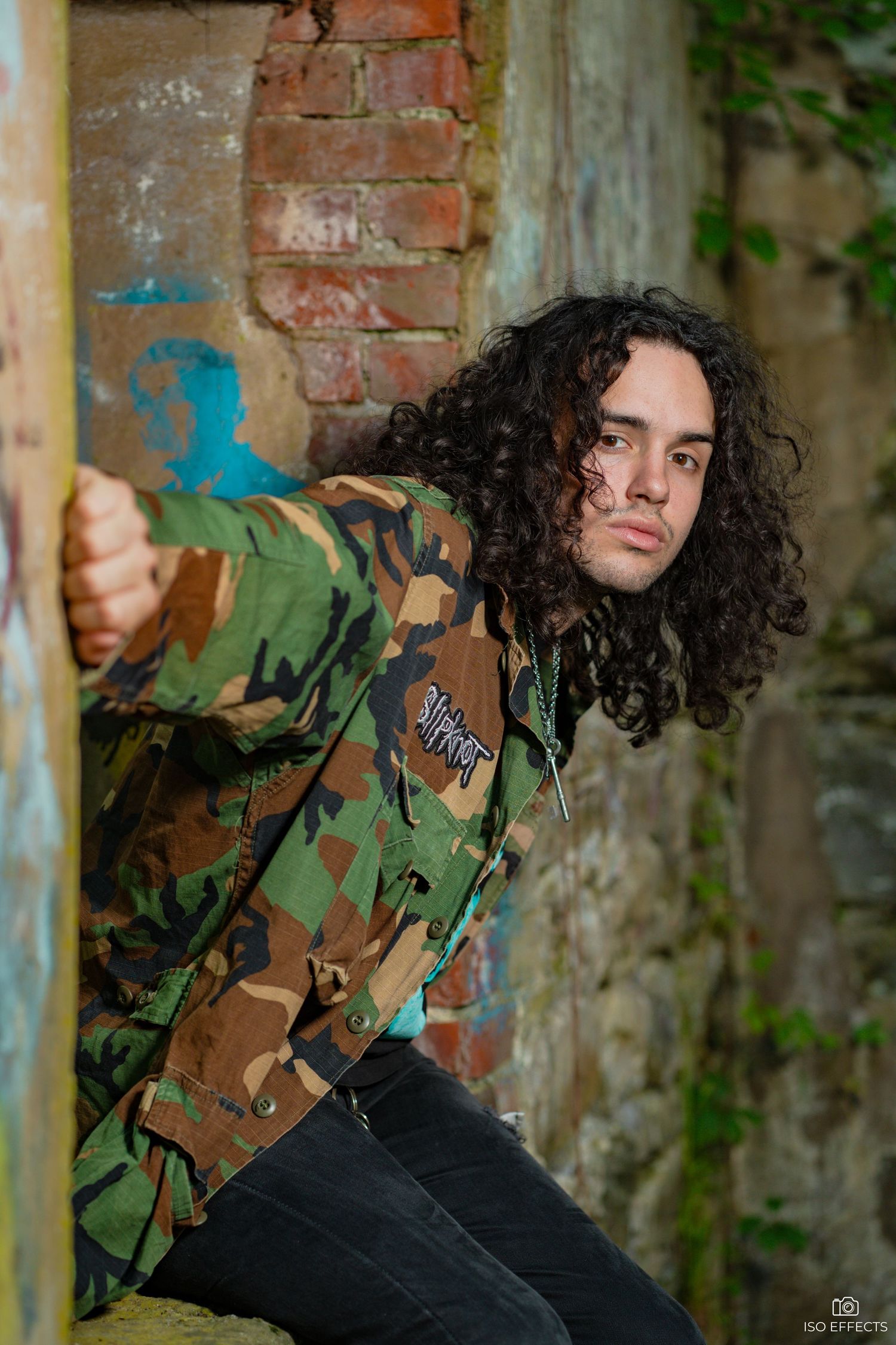 Model in camo jacket poses against graffiti brick wall in urban setting.
