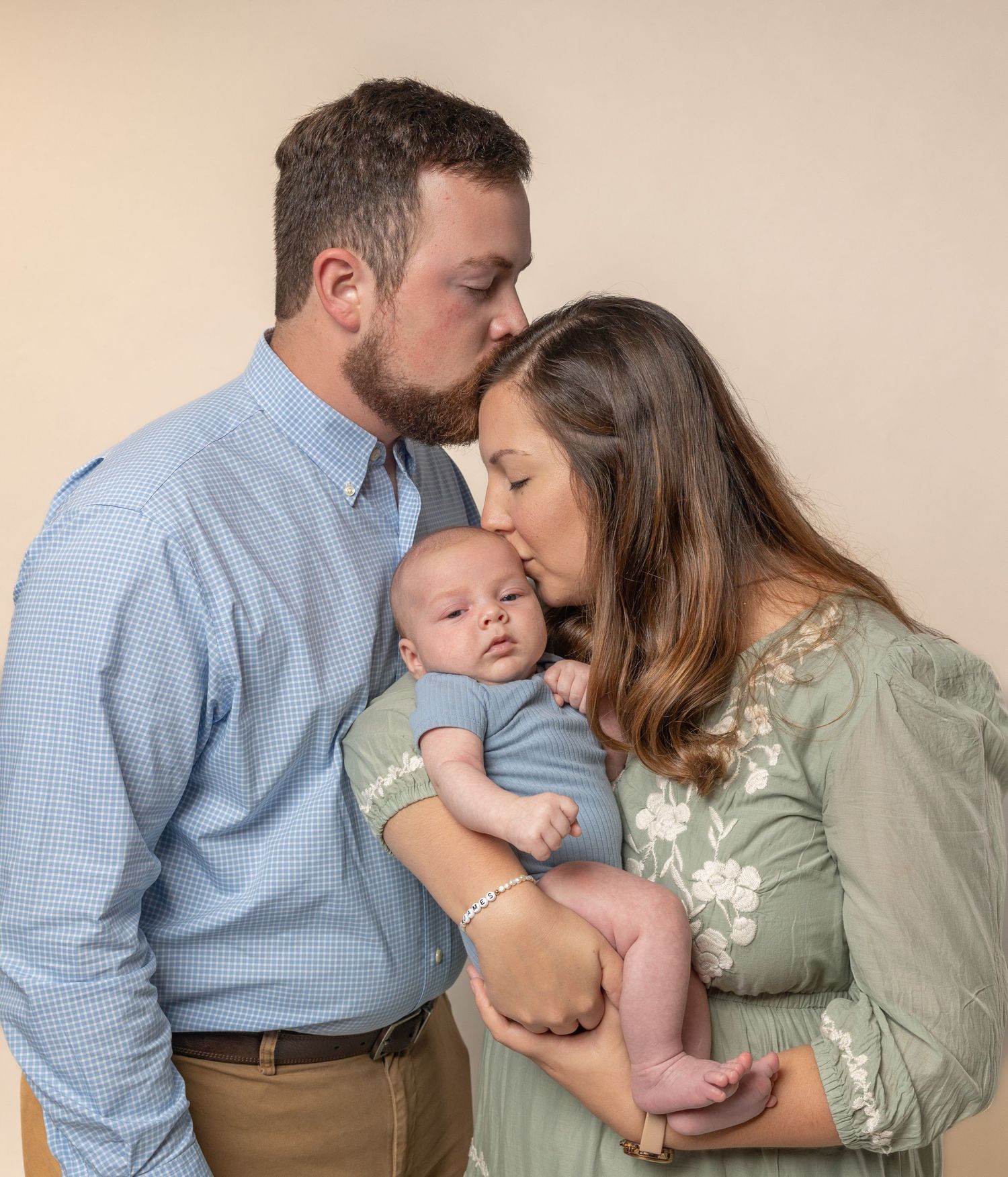 Parents lovingly embrace their newborn baby dressed in blue against a neutral backdrop.