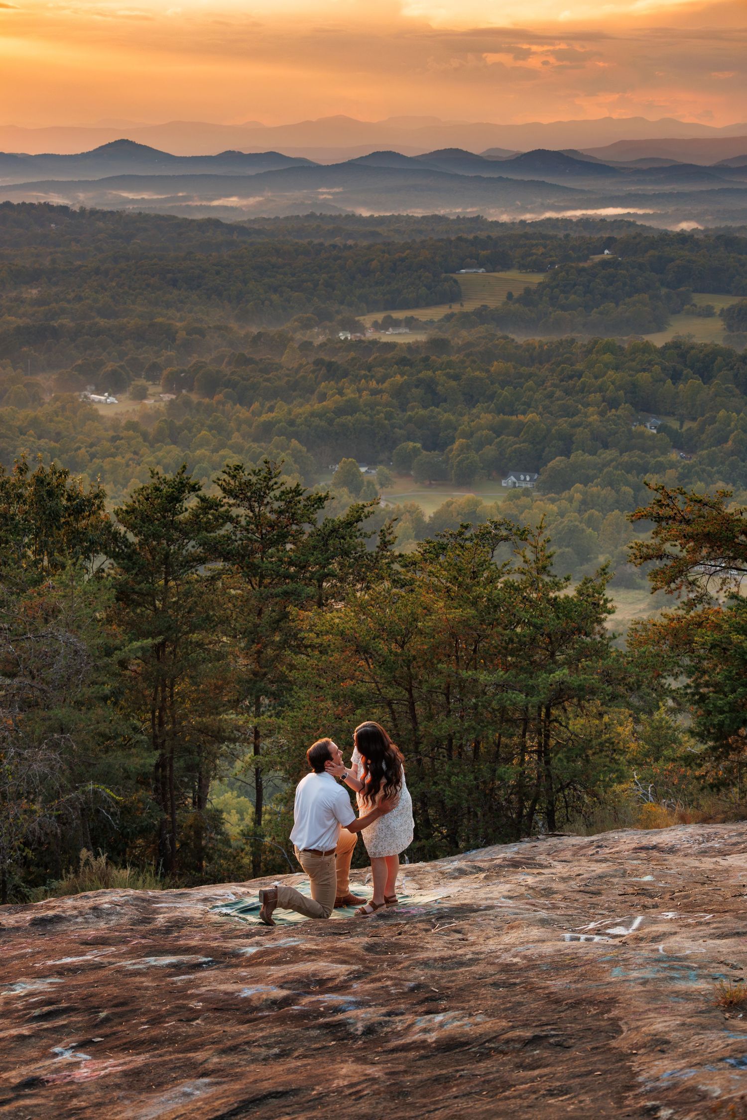 A couple sits together on a rocky mountain overlook at sunset with rolling hills and mountains in the distance.