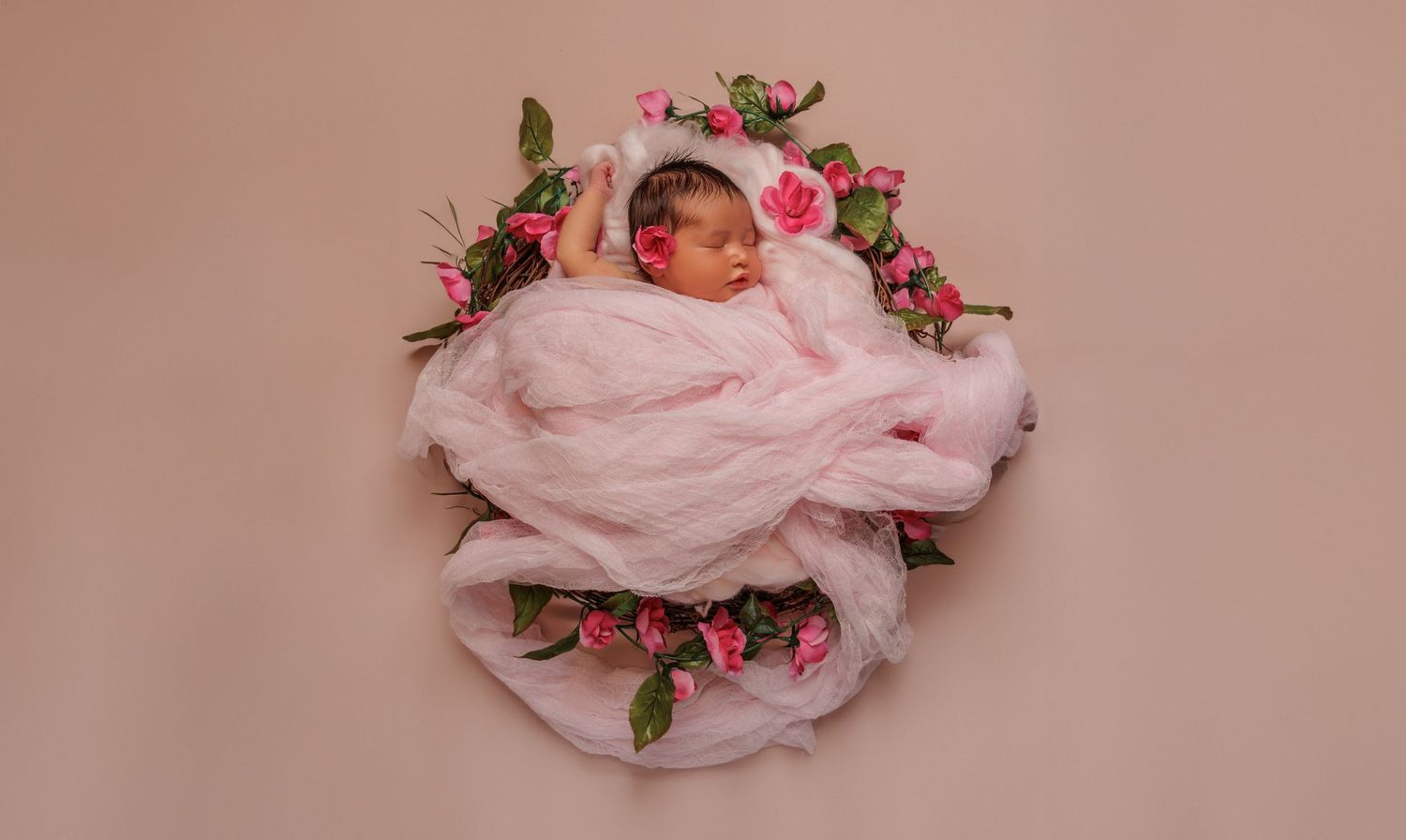 Sleeping newborn baby wrapped in soft pink fabric surrounded by delicate pink roses and floral decorations.