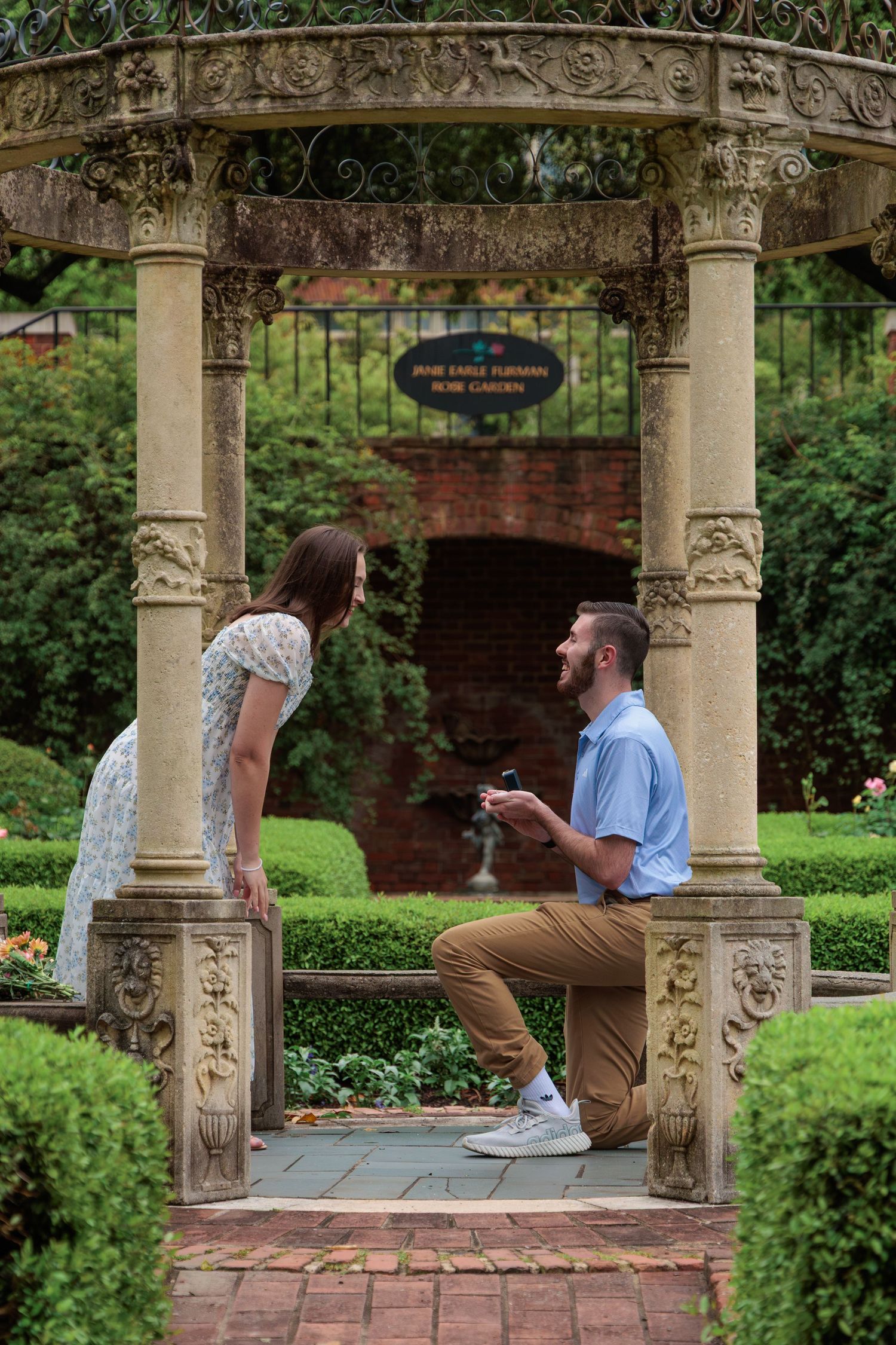 A romantic proposal moment unfolds in a garden gazebo with ornate stone columns and lush greenery.