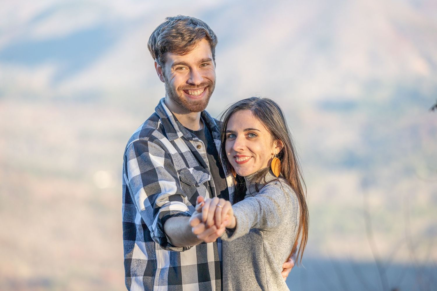 Happy couple in casual attire showing engagement ring outdoors against a blurred mountainous backdrop.