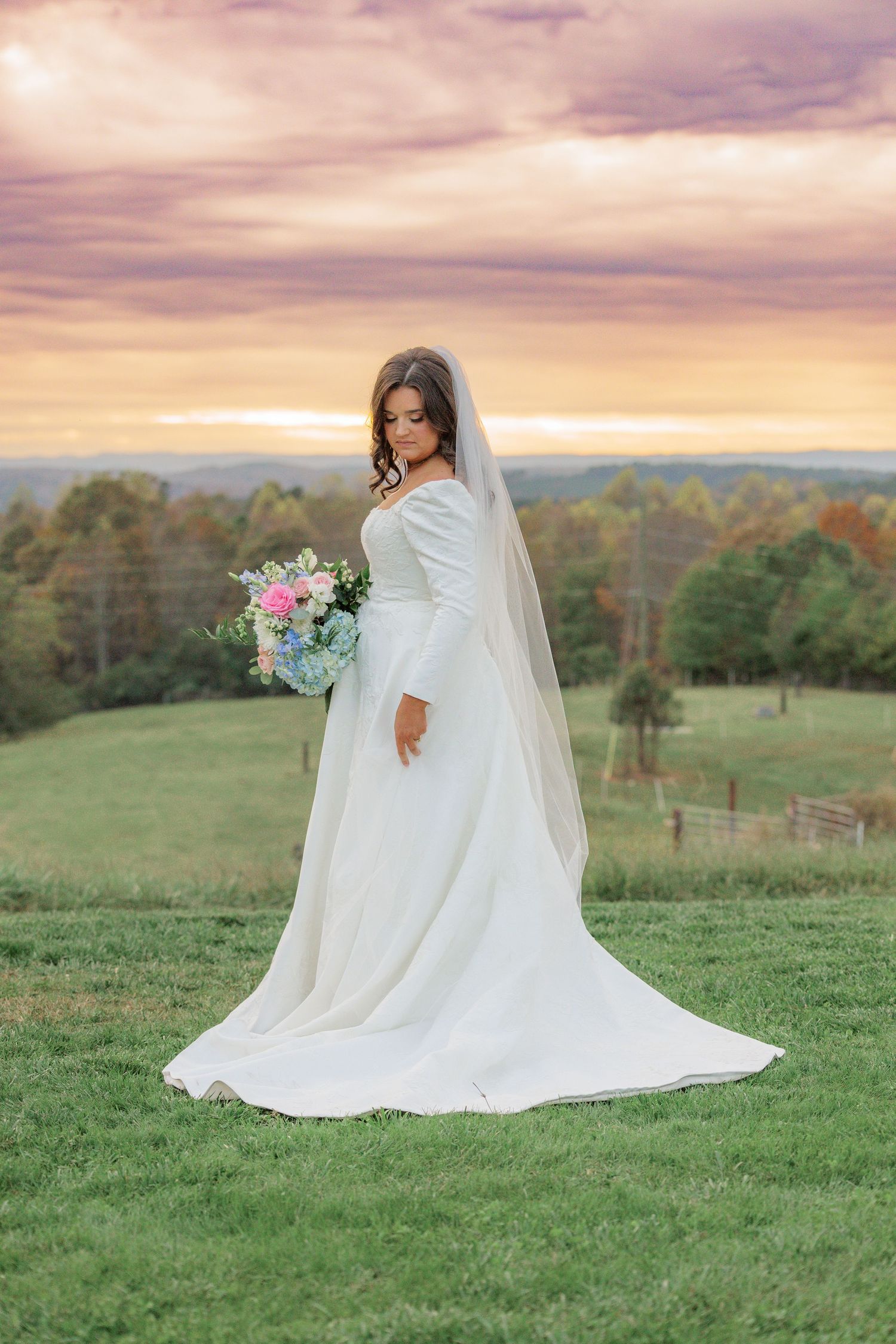 A bride in a flowing white dress and veil poses on a hilltop during sunset with a pink bouquet.