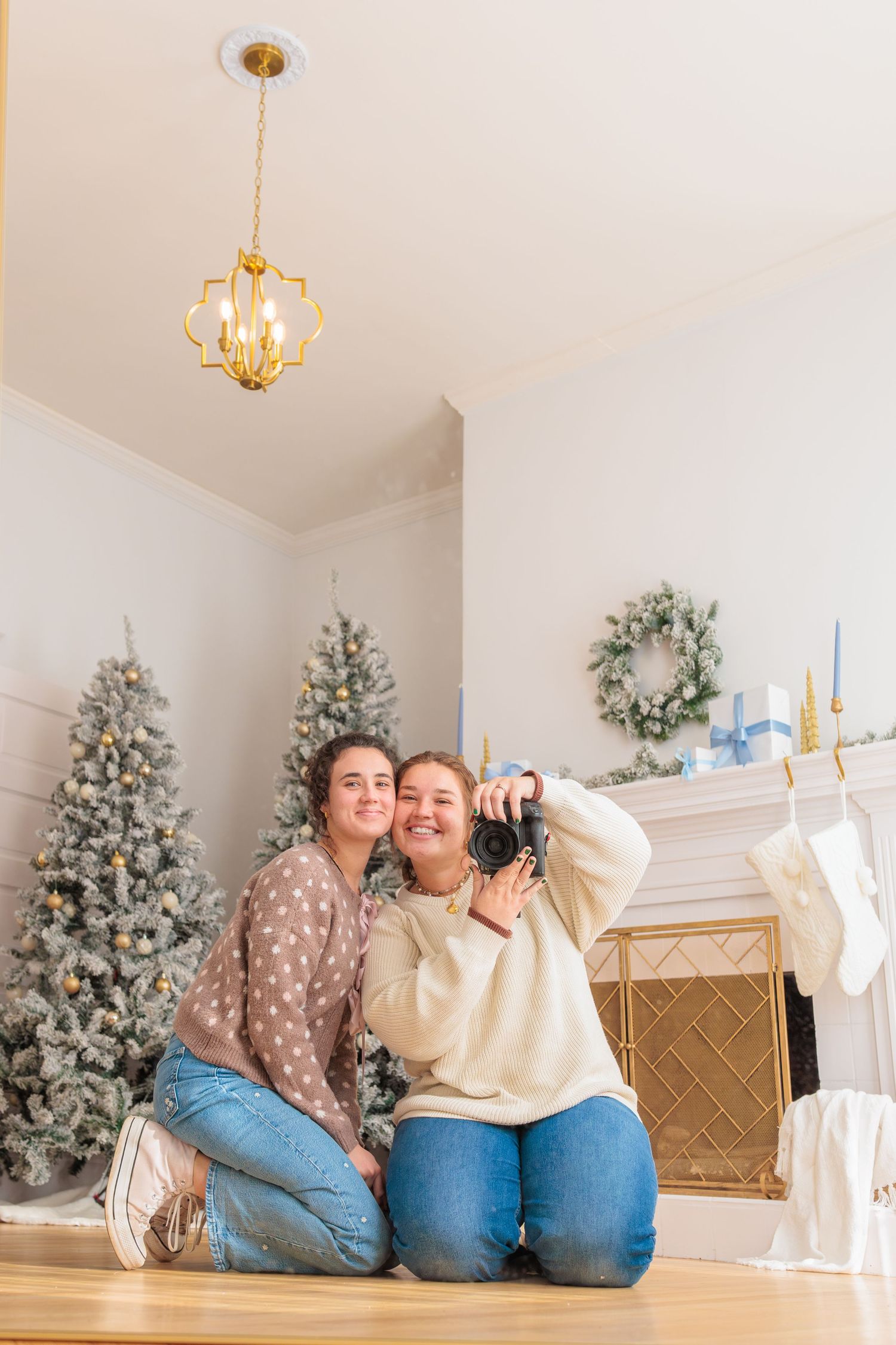 Two people pose in front of decorated Christmas trees and a fireplace with stockings in a cozy holiday setting.