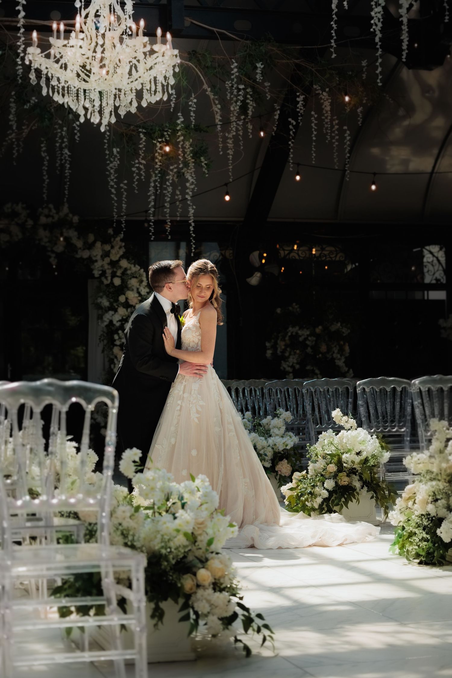 Elegant wedding ceremony setup with crystal chandelier, white chairs, and lush floral arrangements in a romantic evening setting.