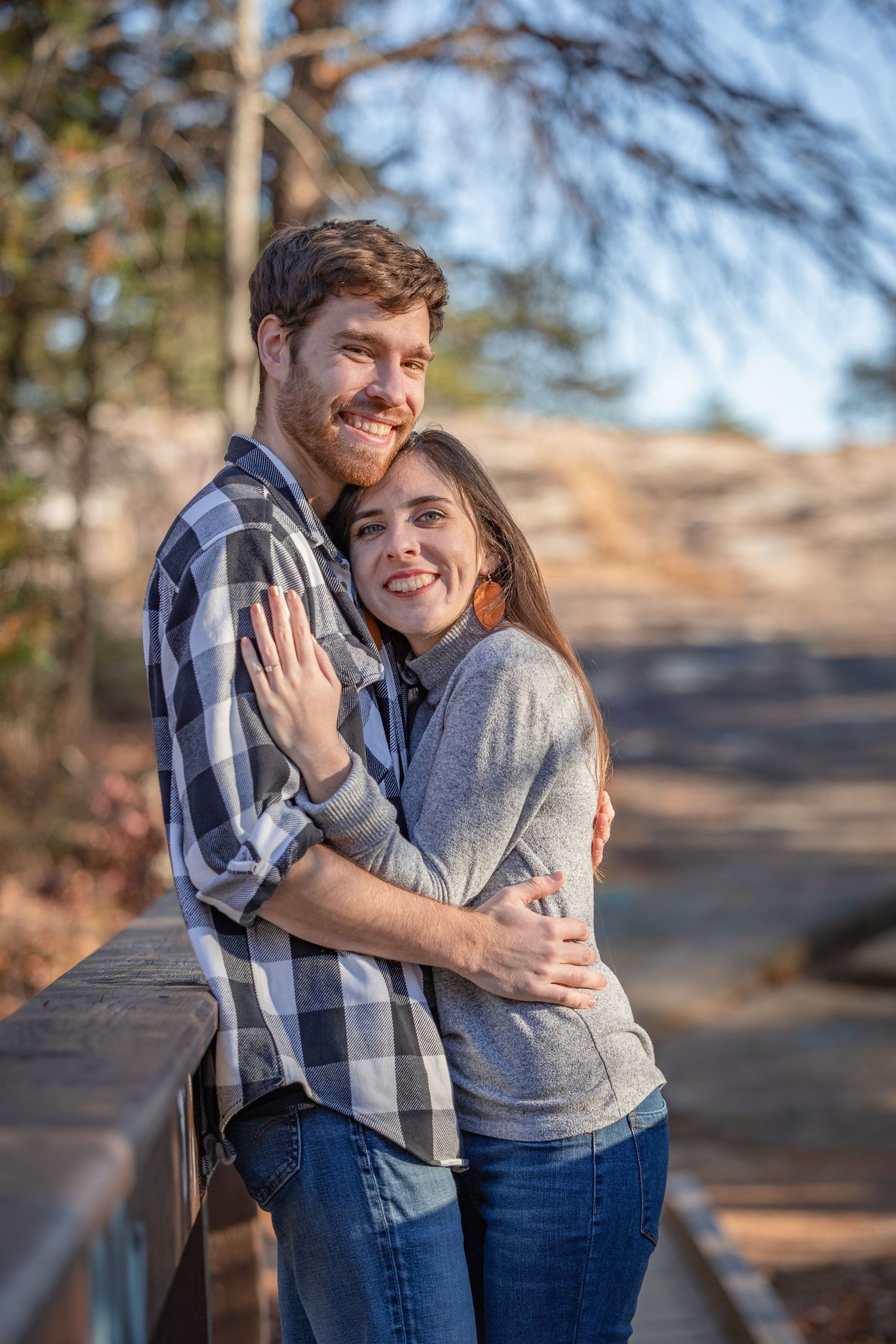 A romantic couple embraces outdoors on a wooden bridge while smiling warmly on an autumn day.